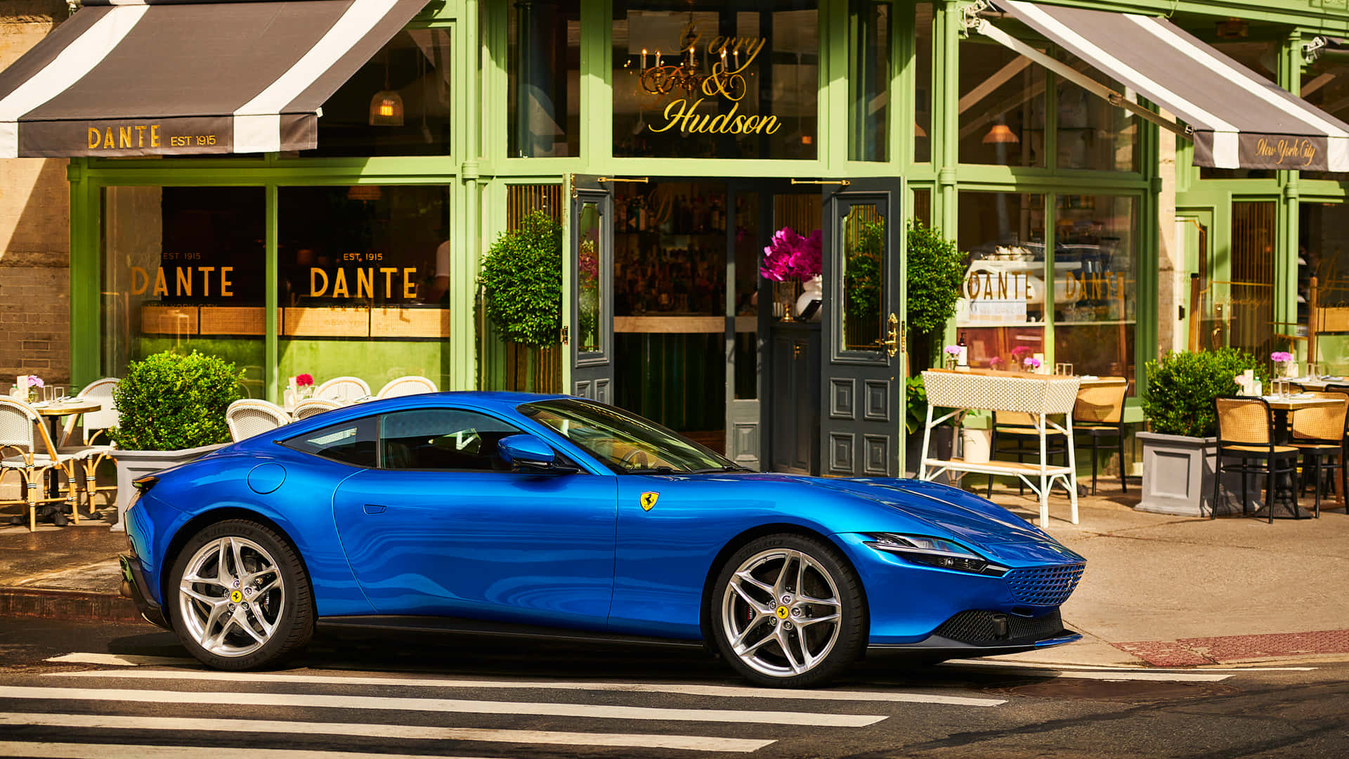 Ferrari Roma 桌布 壁紙 — 一輛鮮藍色的法拉利 Roma 停泊在陽光明媚的街道上，前面是綠色店面，設有戶外咖啡座。