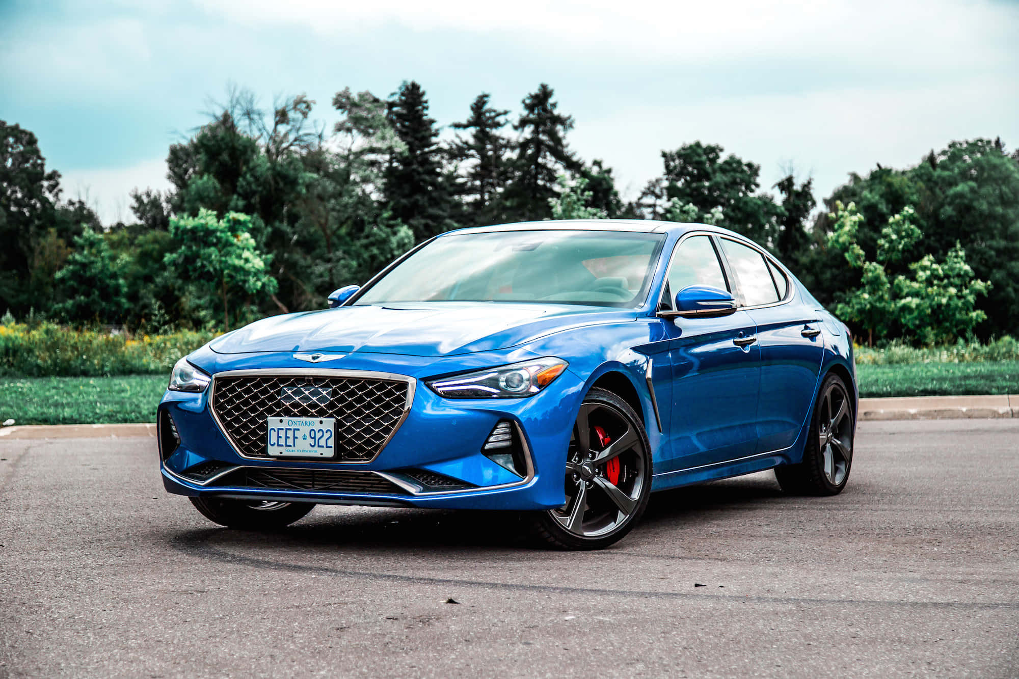 Genesis G70 桌布 壁紙 — 一輛充滿活力的藍色現代 Genesis 轎車，帶有紅色制動卡鉗，停在瀝青路面上，背景有綠樹。