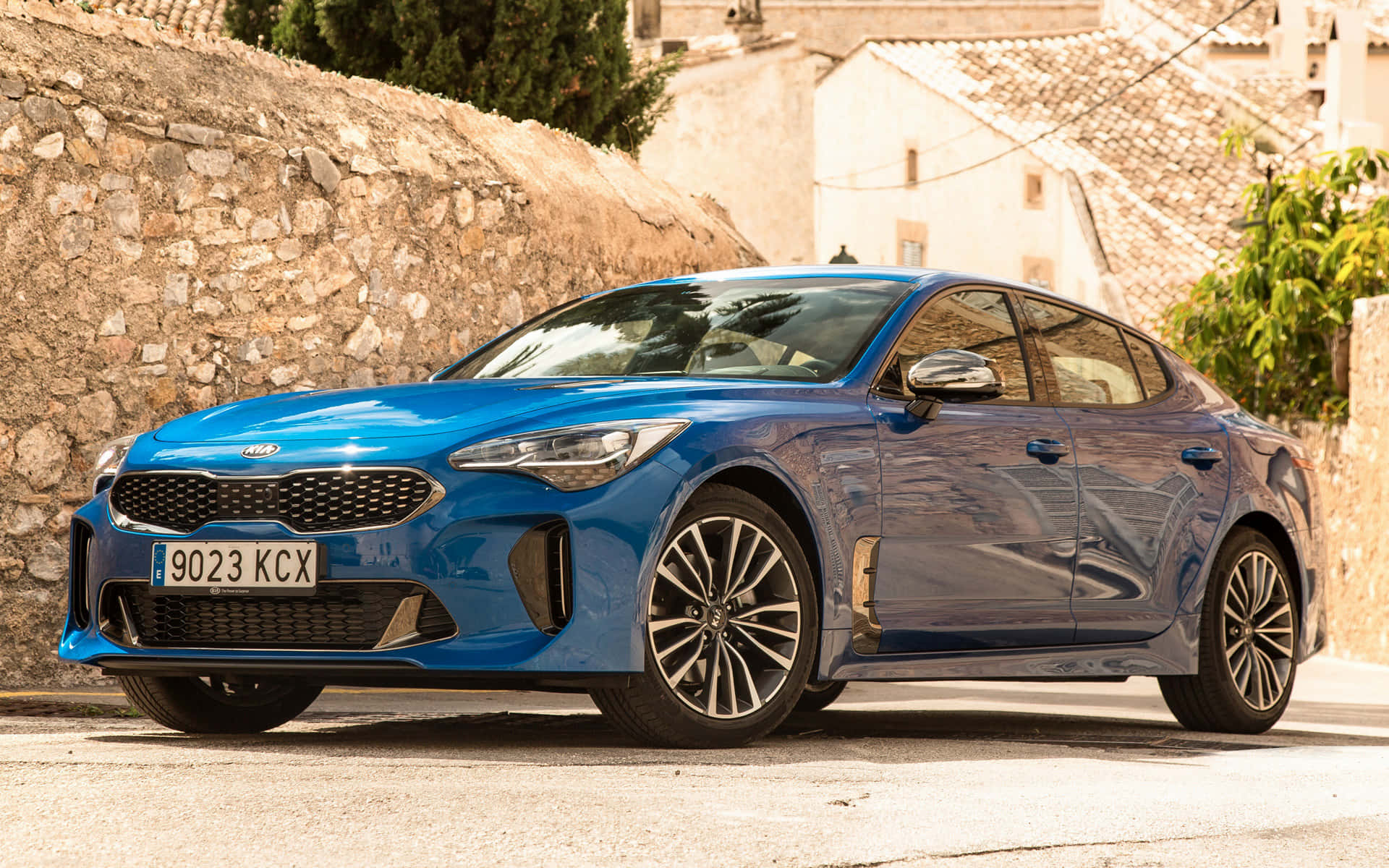 Kia Stinger 桌布 壁紙 — 一輛充滿活力的藍色Kia轎車在明亮的陽光下閃閃發光，停在石砌街道旁，旁邊是質樸的建築。