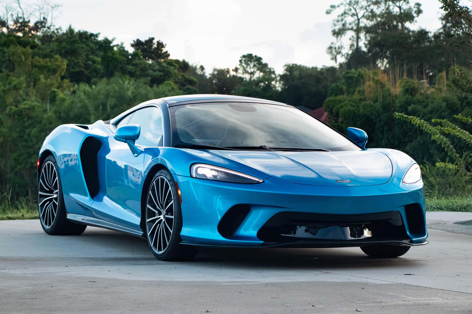 Mclaren Gt 桌布 壁紙 — 一輛充滿活力的藍色McLaren跑車在柔和日光下閃閃發光，停在混凝土表面上，背景有茂密的綠樹。