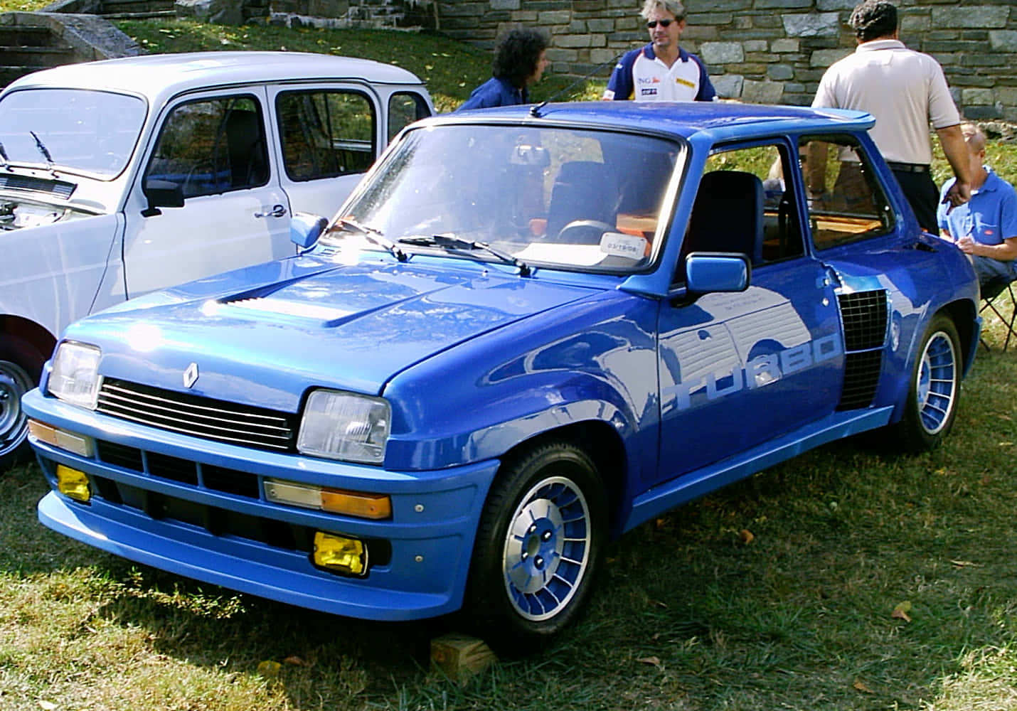 Renault 5 Turbo 桌布 壁紙 — 一輛充滿活力的藍色雷諾5 Turbo跑車在陽光下閃閃發光，停在草地上，附近有人群聚集。