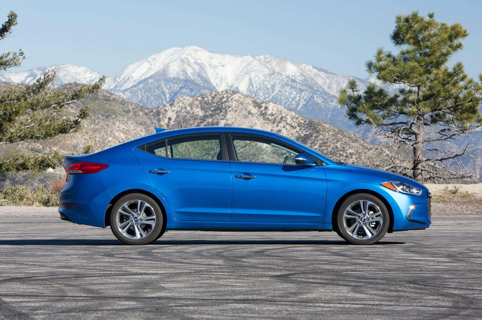 Hyundai Elantra 桌布 壁紙 — 一輛充滿活力的藍色轎車停泊在鋪砌的道路上，背景是白雪覆蓋的山脈和松樹，天空清澈湛藍。