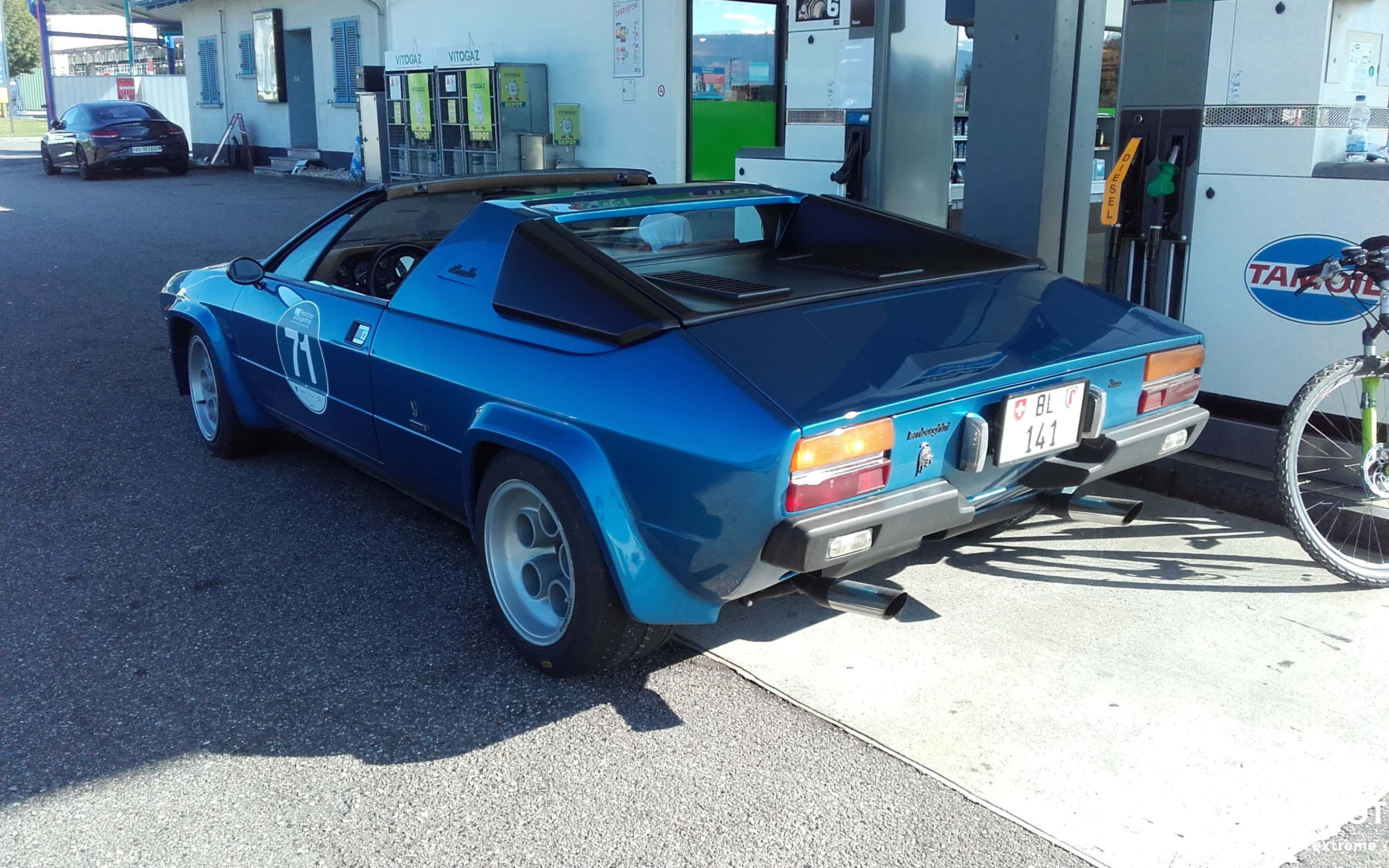 Lamborghini Silhouette 桌布 壁紙 — 一架充滿活力的藍色復古林寶堅尼Jalpa開篷車停在加油站，附近有一輛單車。