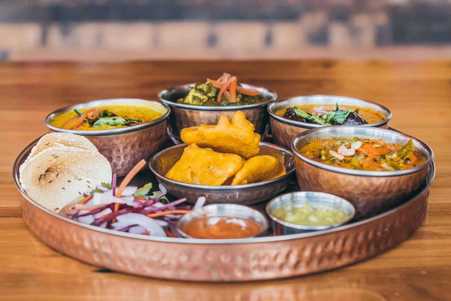 Thali 桌布 壁紙 — 一個充滿活力的印度塔利餐，盛放在錘紋銅托盤上，配有色彩繽紛的咖喱、薩莫薩三角餃和烤餅，置於木桌上。
