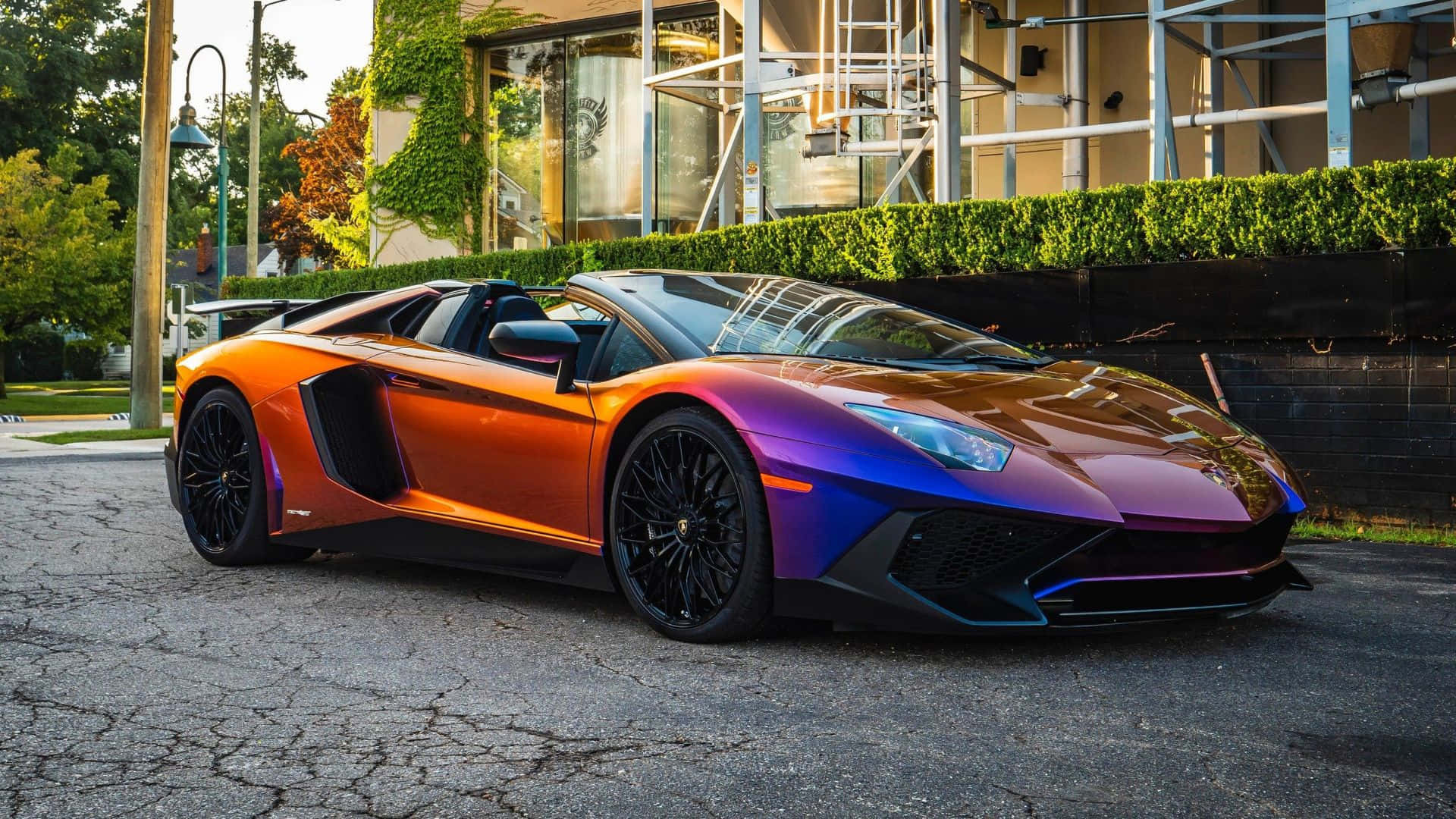  壁紙 — 一輛充滿活力的彩虹色林寶堅尼Aventador開篷跑車停泊在裂開的瀝青路上，旁邊有綠色籬笆和現代建築。