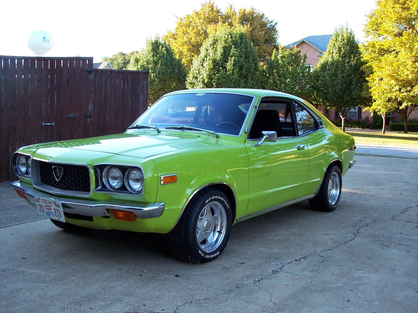 Mazda Rx-3 桌布 壁紙 — 一輛充滿活力的青檸綠色經典雙門跑車停在裂開的混凝土車道上，背景有樹木和木柵欄。