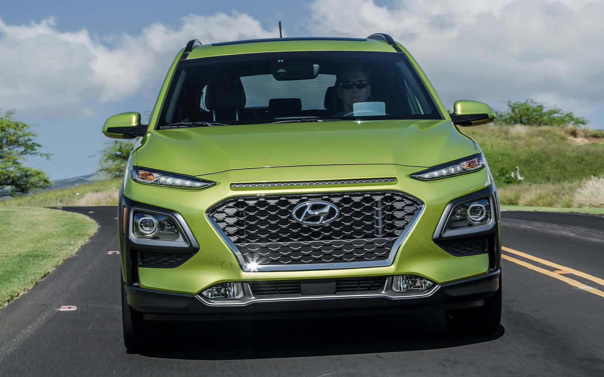 Hyundai Kona 桌布 壁紙 — 一輛充滿活力的青檸綠色現代SUV在部分多雲的天空下沿著蜿蜒的道路正面行駛。