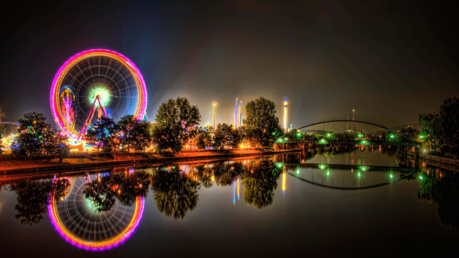  壁紙 — 一個充滿活力的夜景，特色是色彩繽紛的摩天輪和橋樑，倒映在平靜的水面上，周圍環繞著樹木。