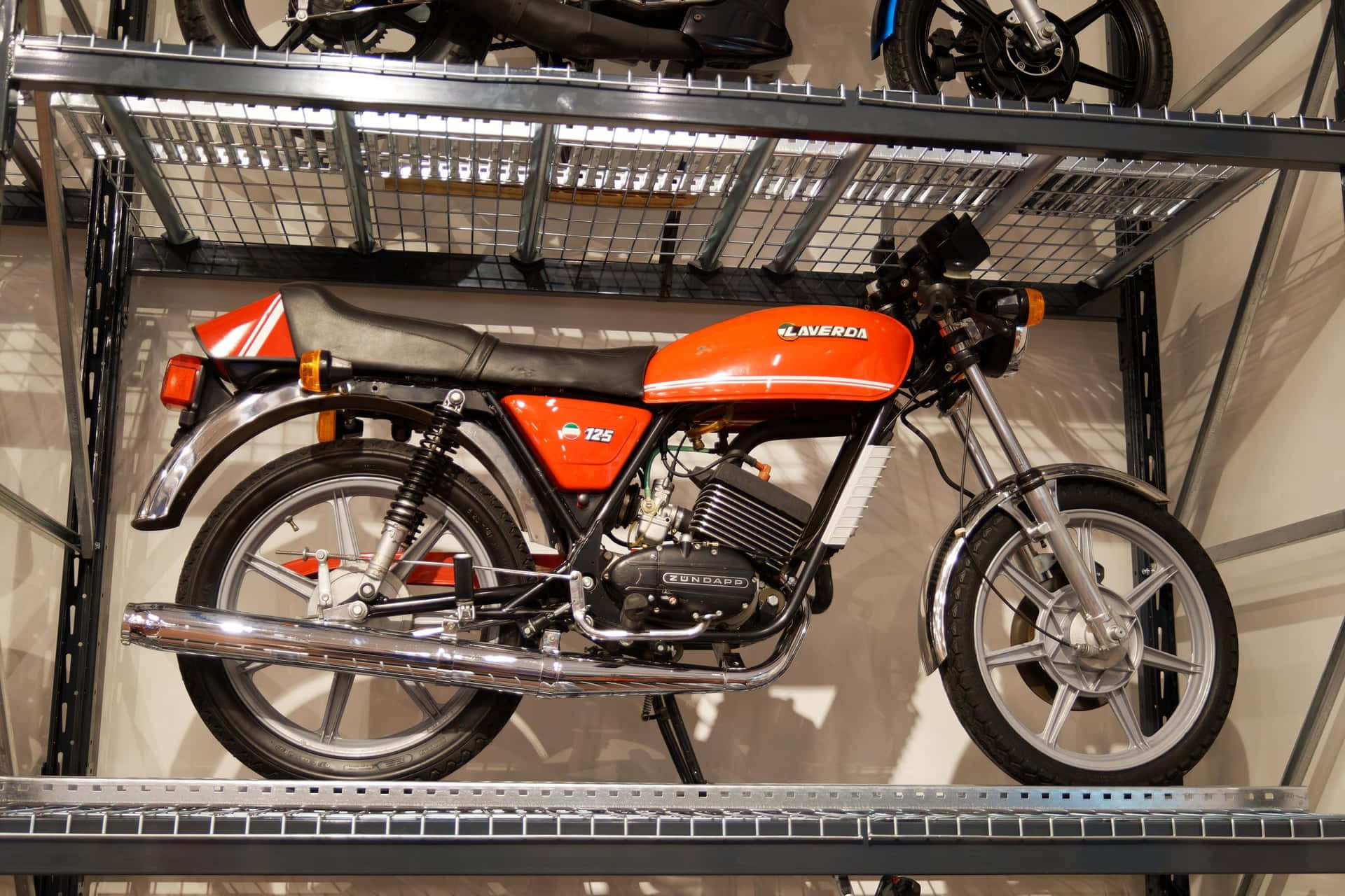  壁紙 — 一輛鮮豔的橙色Laverda 125電單車展示在光線充足的室內車庫的金屬架上。