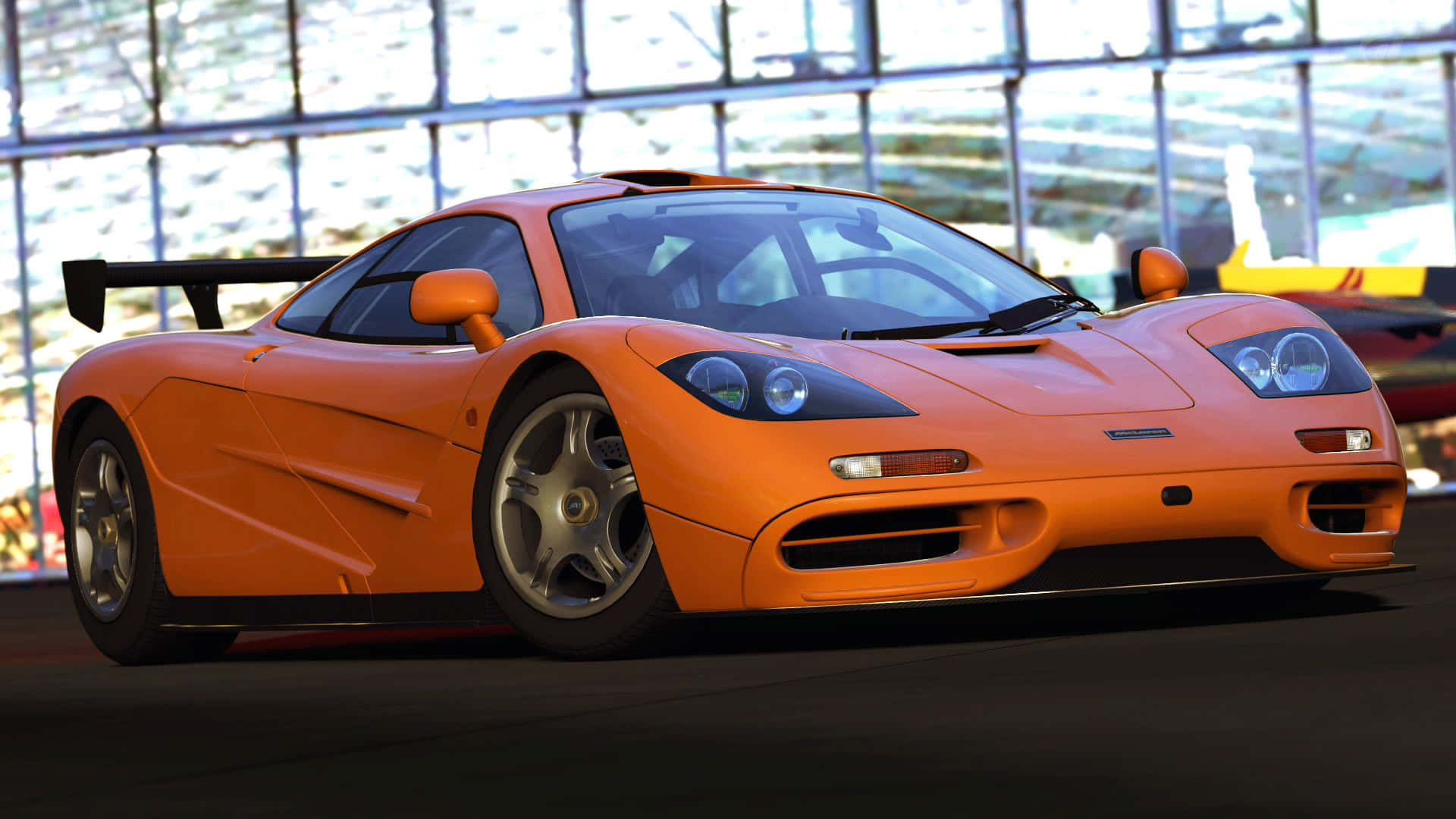 Mclaren F1 桌布 壁紙 — 一輛充滿活力的橙色McLaren F1超級跑車在現代玻璃牆車庫內，在明亮燈光下閃閃發光。