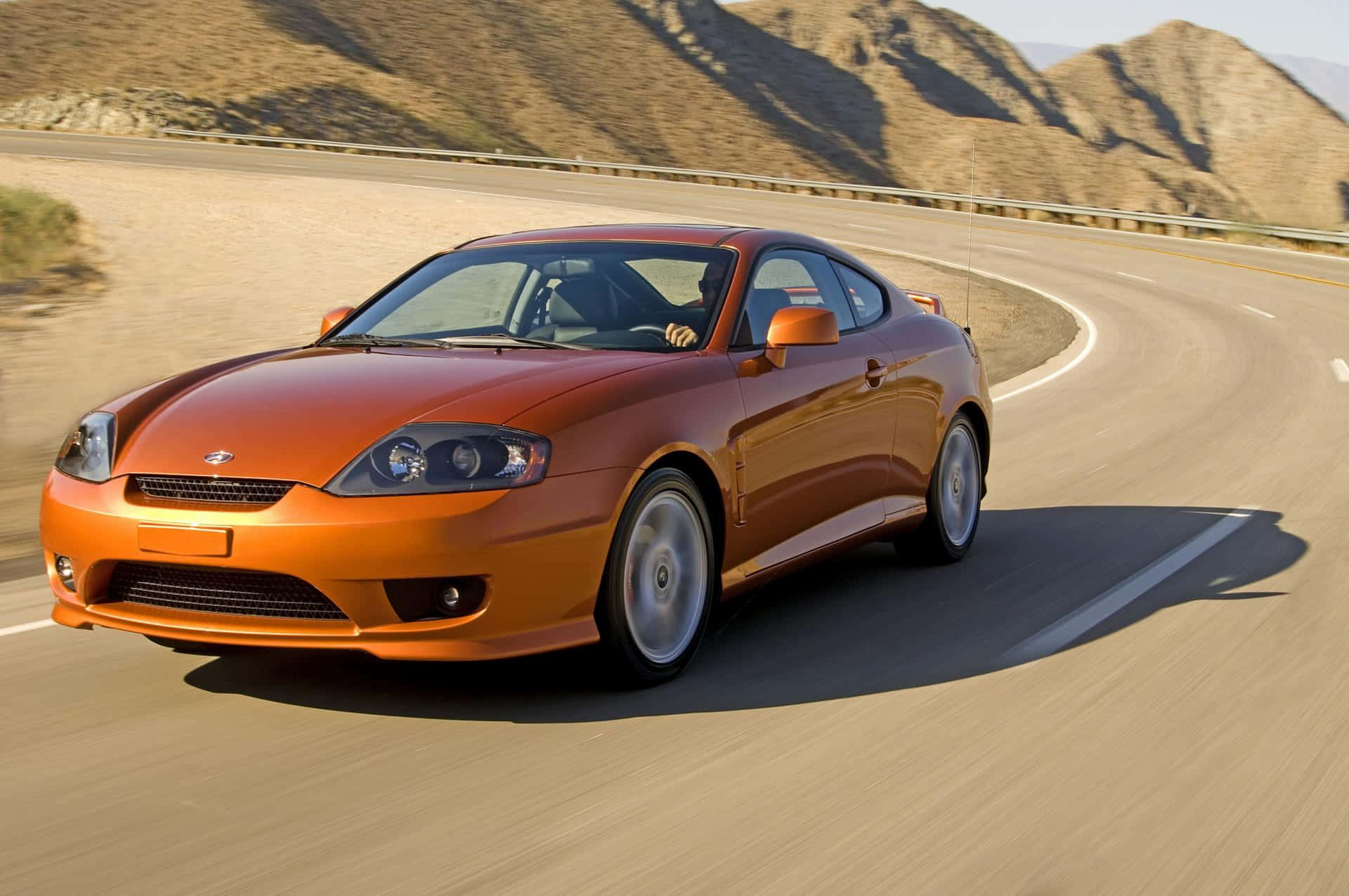 Hyundai Tiburon 桌布 壁紙 — 一輛充滿活力的橙色跑車在蜿蜒的沙漠公路上疾馳，背景是崎嶇的山脈。