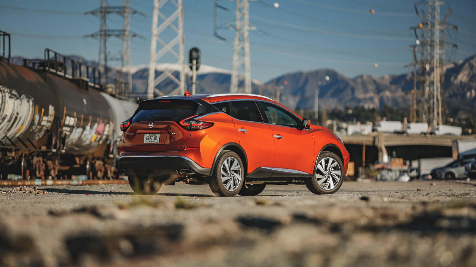Nissan Murano 桌布 壁紙 — 一輛充滿活力的橙色SUV停泊在火車車廂附近，背景有山脈和電線，天空清澈湛藍。