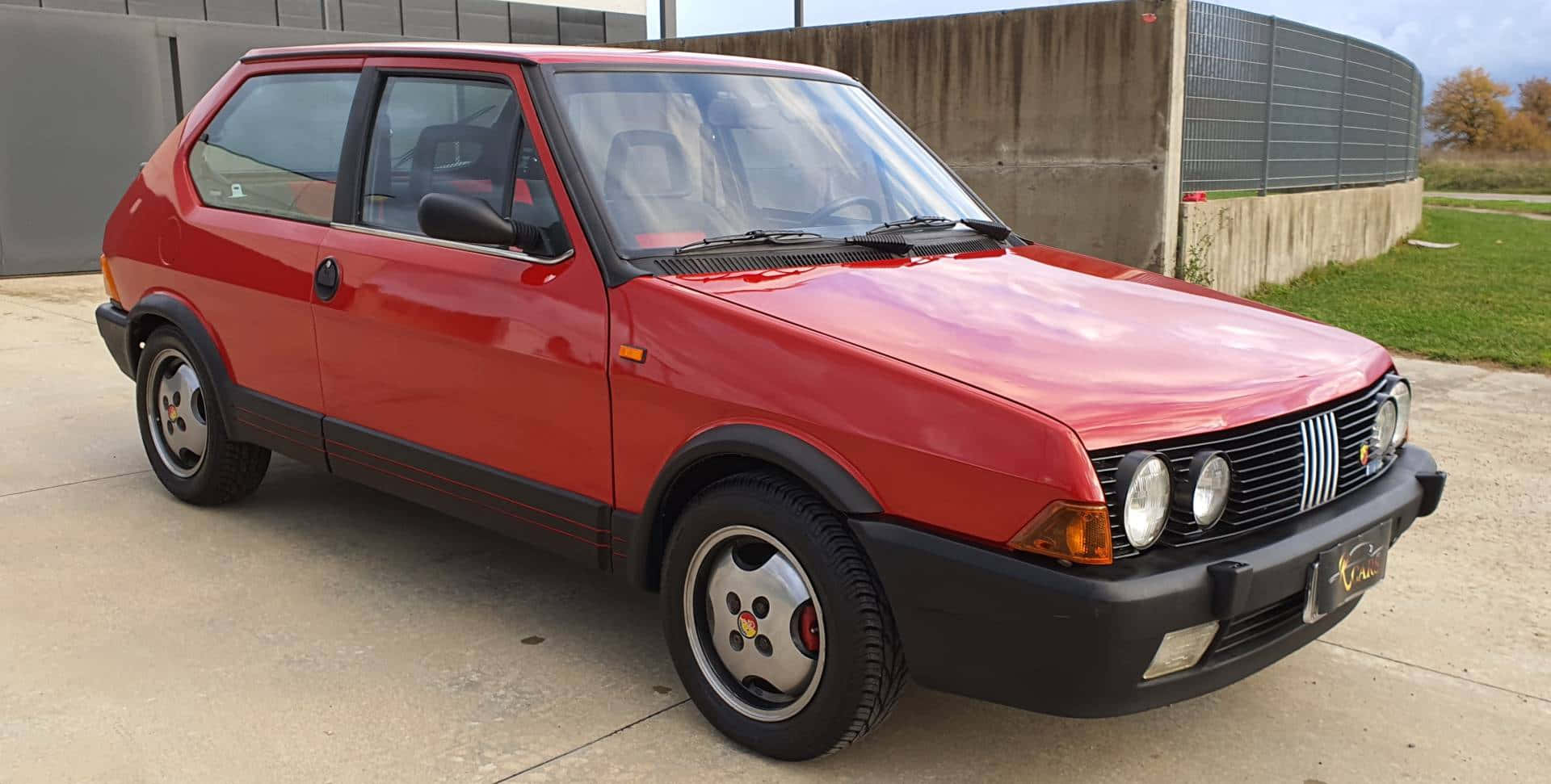 Fiat Ritmo 桌布 壁紙 — 一輛鮮紅色的經典菲亞特Uno停泊在混凝土空地旁，旁邊有草地和混凝土牆。