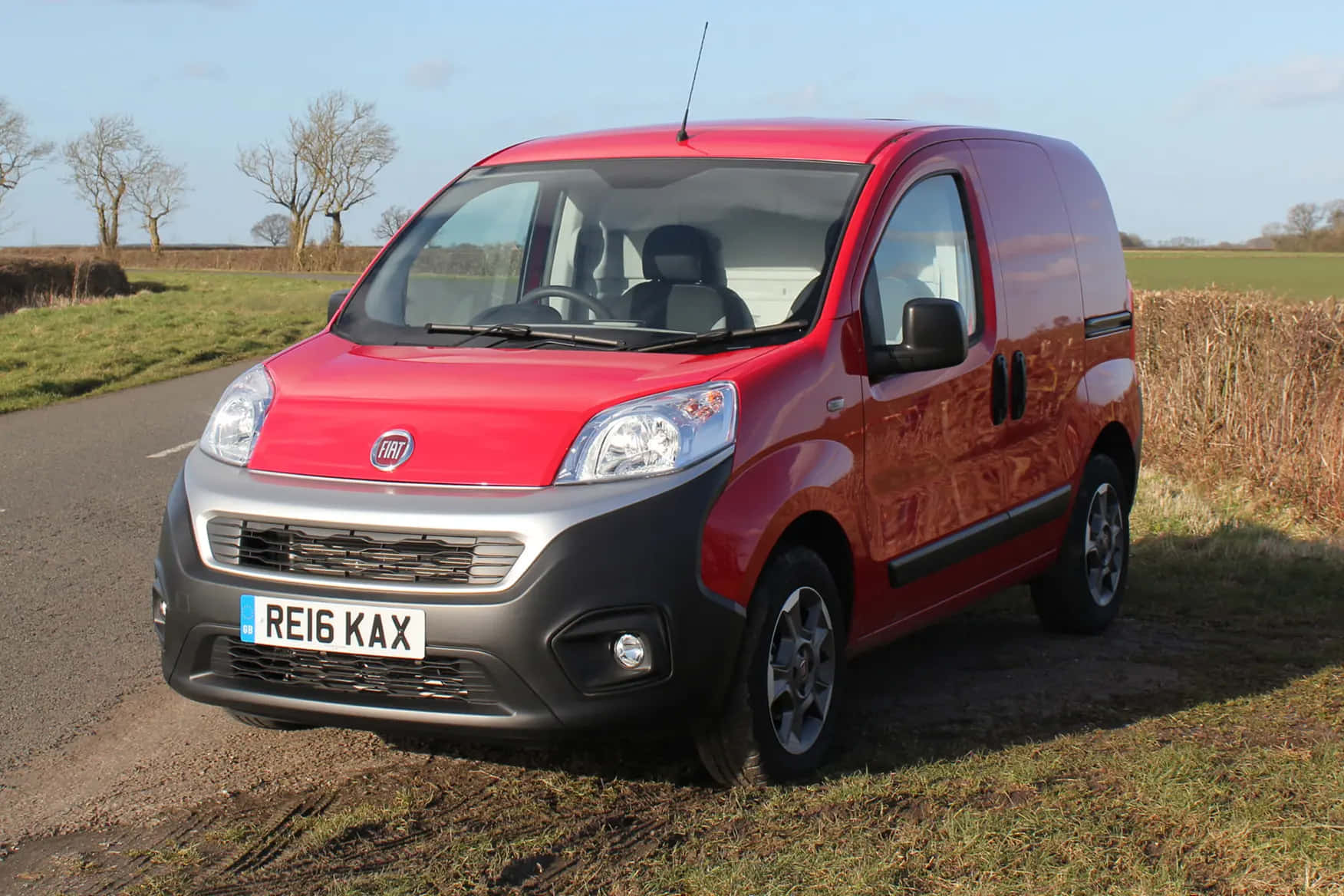 Fiat Fiorino 桌布 壁紙 — 一架鮮紅色嘅Fiat貨車停喺鄉郊路邊，周圍有光禿禿嘅樹木同開闊嘅田野，上面係晴朗嘅藍天。