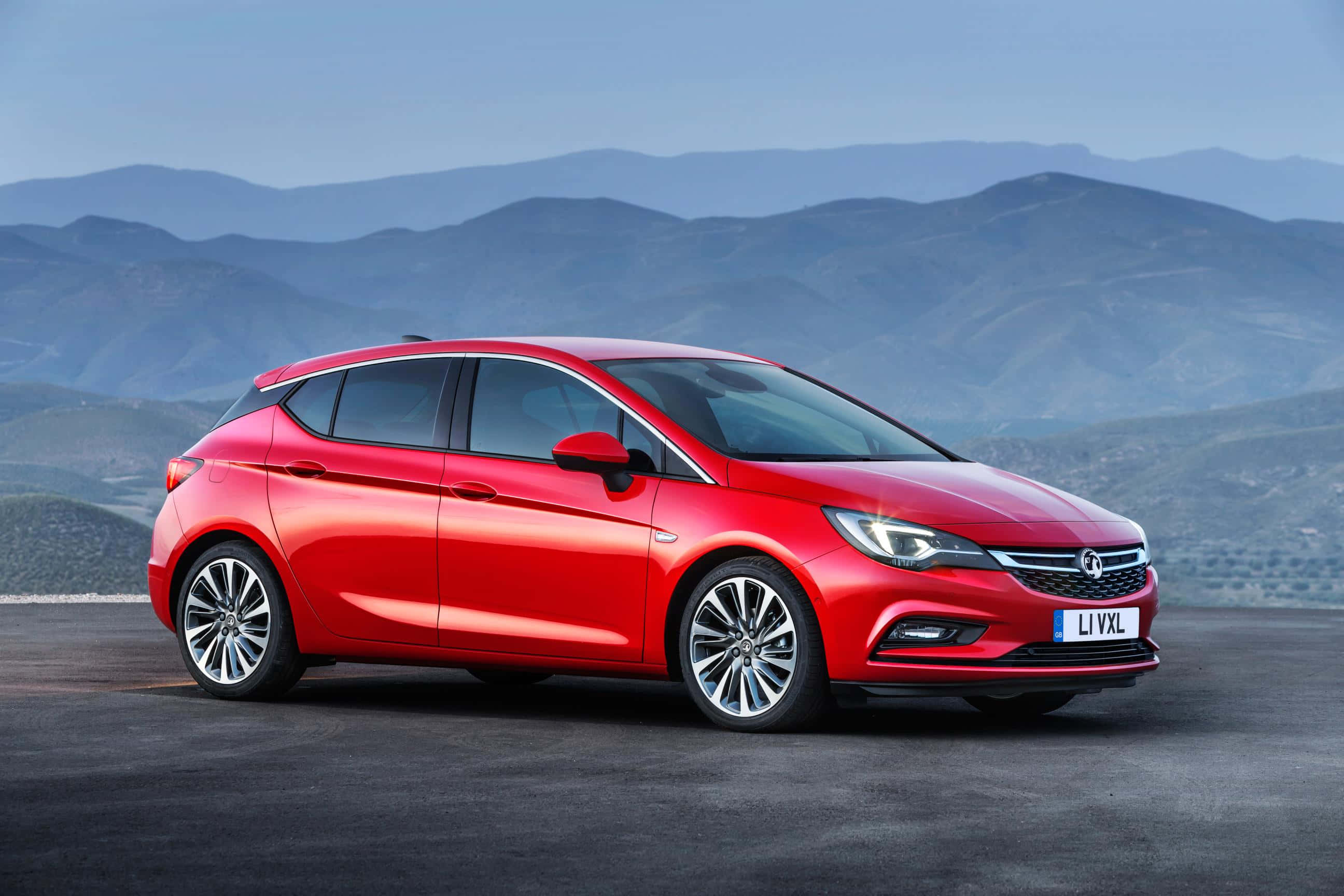 Opel Astra 桌布 壁紙 — 一輛鮮紅色的掀背車在日光下閃閃發光，背景是層疊的山脈。