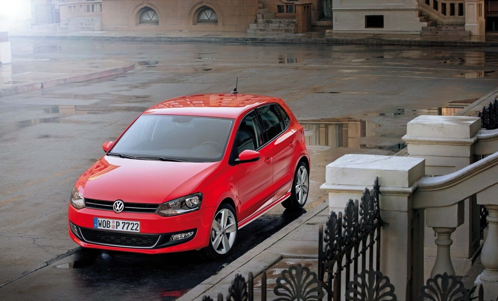 Volkswagen Polo 桌布 壁紙 — 一輛鮮紅色的福士Polo停泊在濕漉漉的城市街道上，映照著多雲的天空和石頭建築。