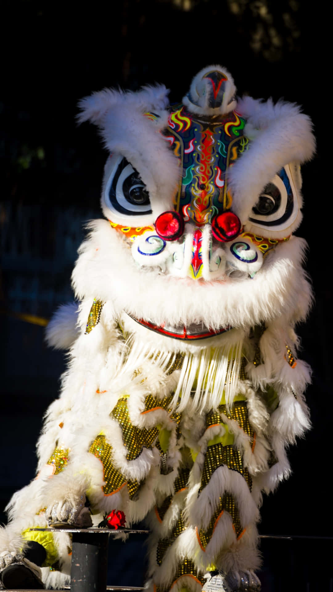  壁紙 — 一件色彩繽紛、繡有圖案和紅色絨球嘅白色舞獅服裝，喺黑暗背景下被照亮。