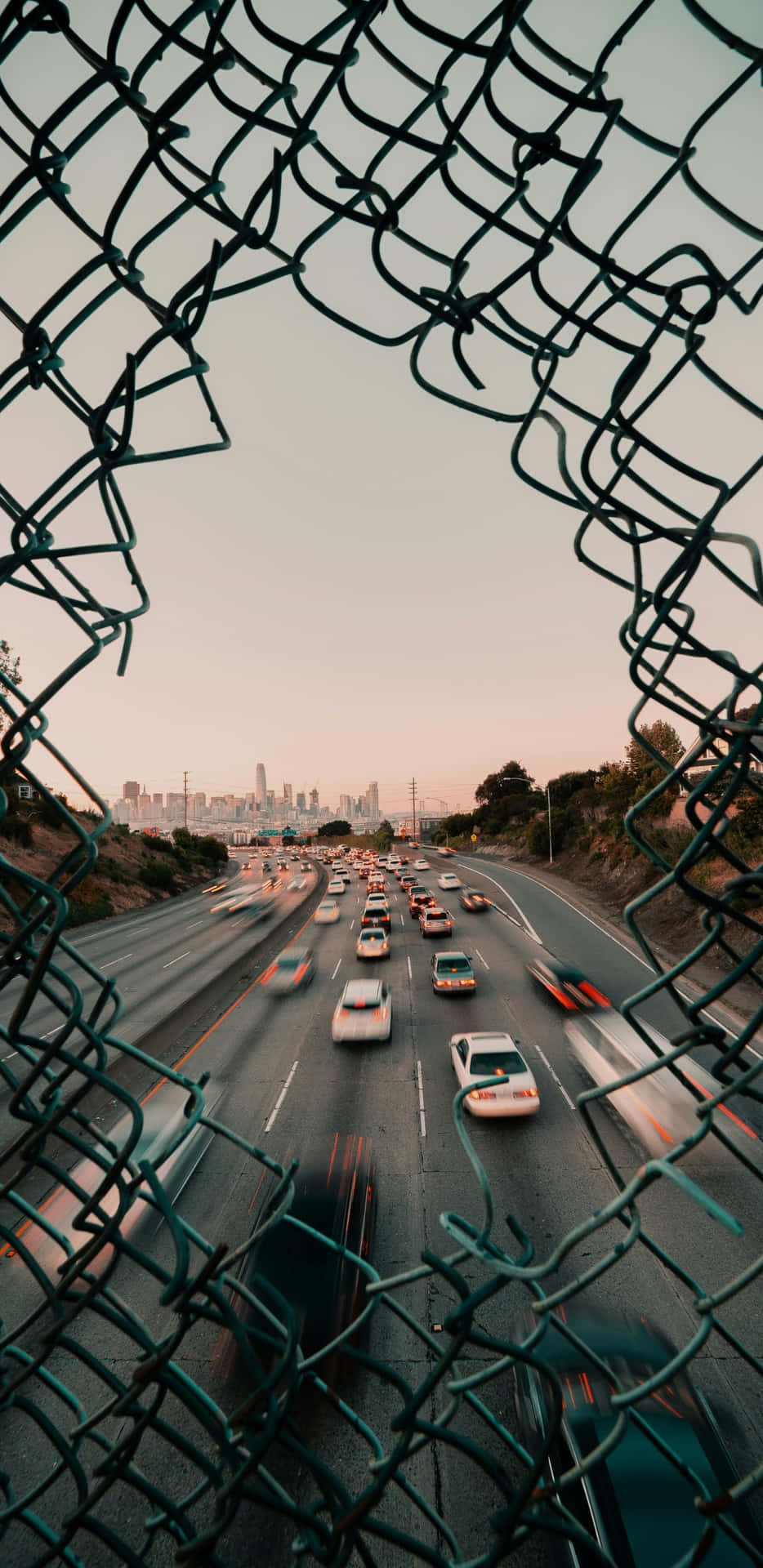  壁紙 — 透過鐵絲網圍欄看到的景色顯示出模糊的汽車在高速公路上疾馳，朝向日落時分的城市天際線。