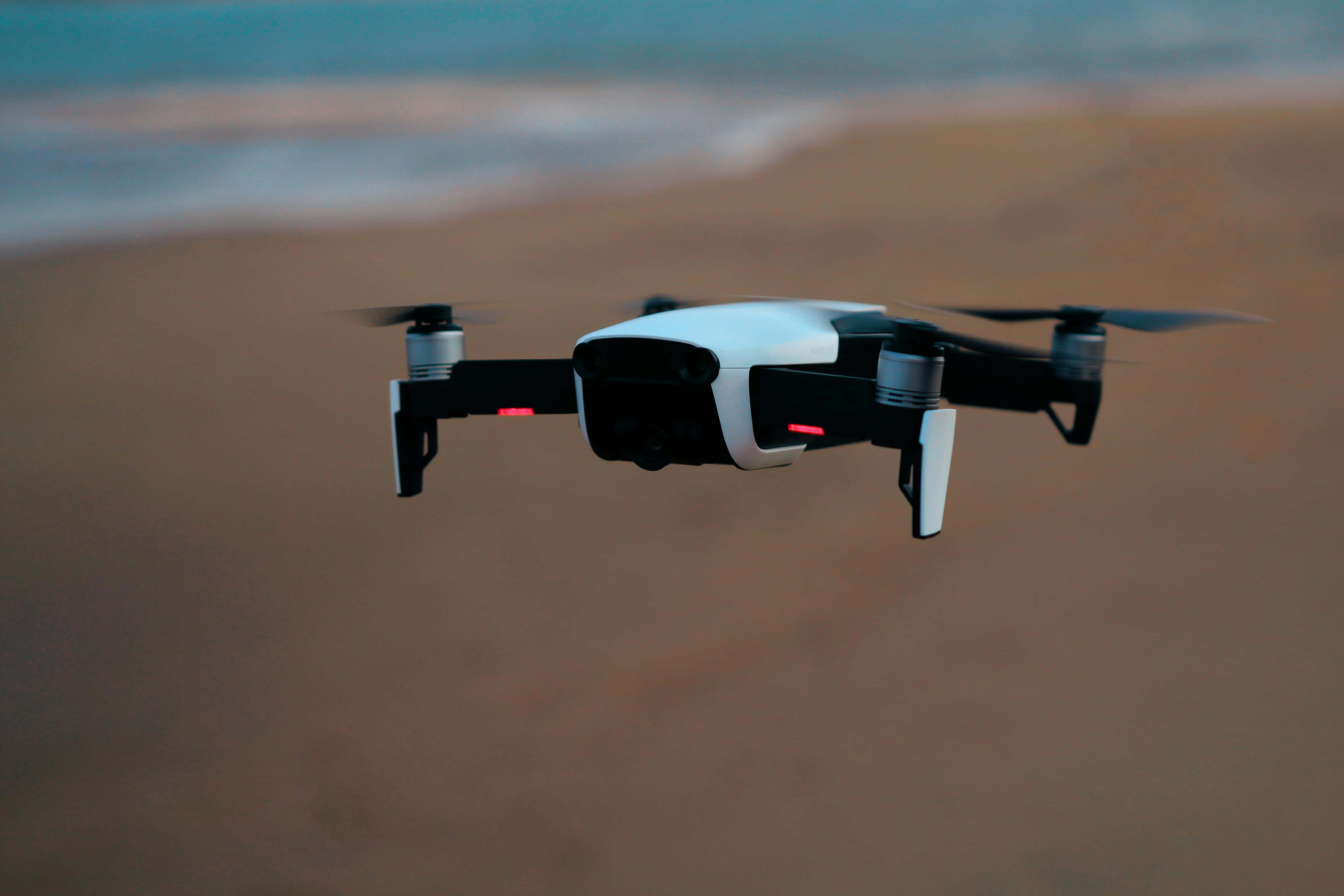 Drone Camera 桌布 壁紙 — 一架白色和黑色的無人機在黃昏時分盤旋在沙灘上空，背景是模糊的海浪。