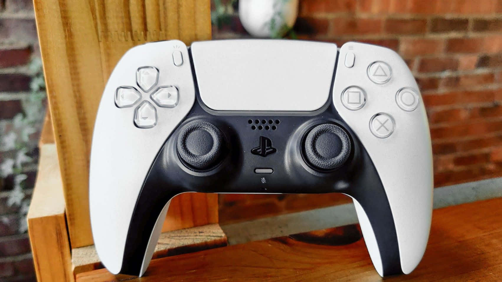  壁紙 — 一個白色和黑色的PlayStation 5 DualSense控制器放在木質表面上，背景是磚牆。