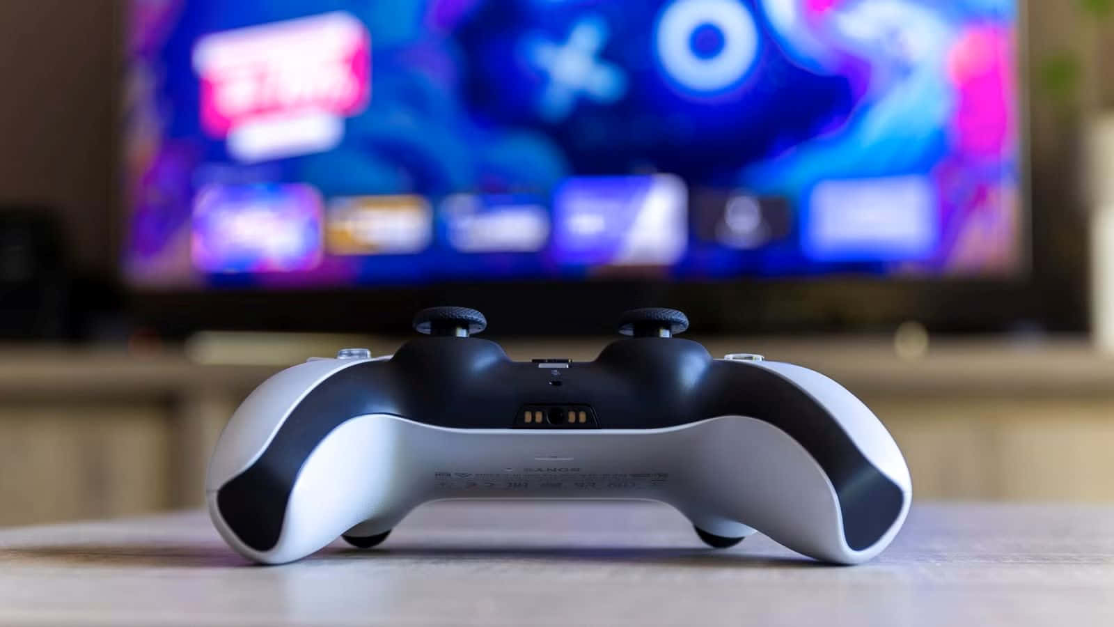  壁紙 — 一個白色和黑色的PlayStation控制器放在木桌上，背景是色彩繽紛且模糊的電視屏幕。