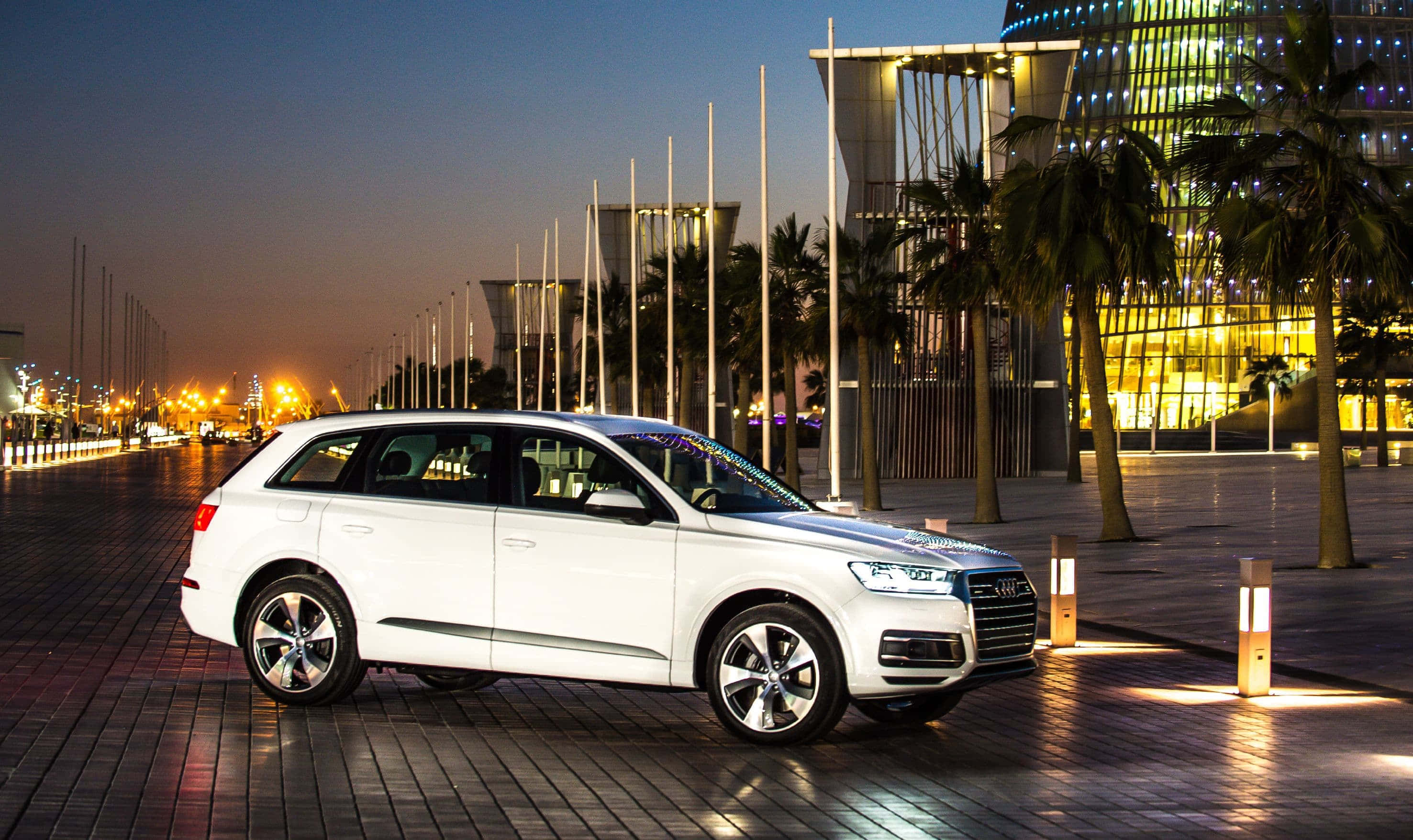 Audi Q7 桌布 壁紙 — 一輛白色奧迪SUV在暮色燈光下，在鋪砌的廣場上閃閃發光，旁邊是現代建築和棕櫚樹。