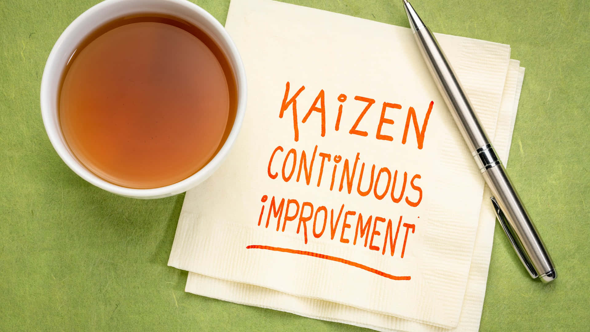  壁紙 — 一杯白色茶放在一張餐巾旁邊，餐巾上用橙色記號筆寫著「Kaizen Continuous Improvement」，旁邊有一支銀色筆在綠色表面上。