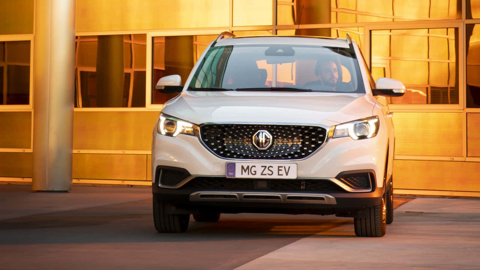  壁紙 — 一輛白色MG ZS EV SUV停在一棟金色玻璃建築前，溫暖的日落燈光。