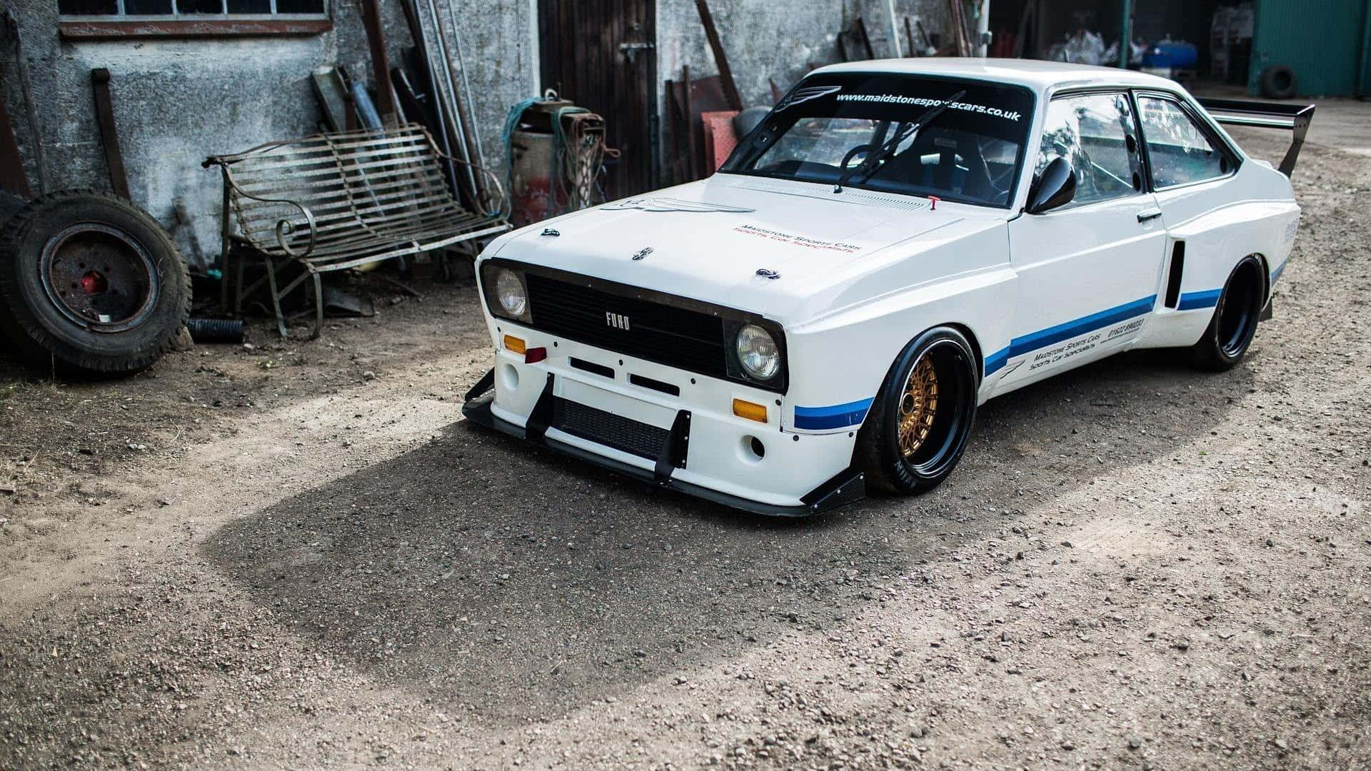 Ford Escort 桌布 壁紙 — 一架白色改裝嘅Fiat拉力賽車，帶有藍色條紋同金色輪圈，停泊喺一個鄉村風格車庫庭院嘅碎石地上。