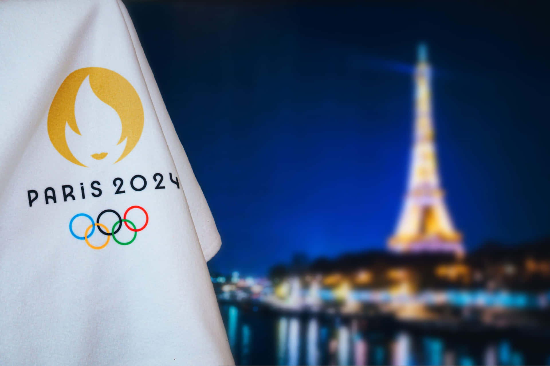  壁紙 — 前景中一件白色巴黎2024奧運襯衫帶有標誌，背景中模糊的艾菲爾鐵塔在夜晚發光。