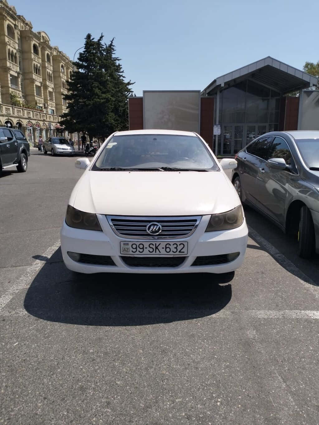  壁紙 — 一輛白色轎車停泊在陽光明媚的城市地段，附近有歷史建築和其他汽車。