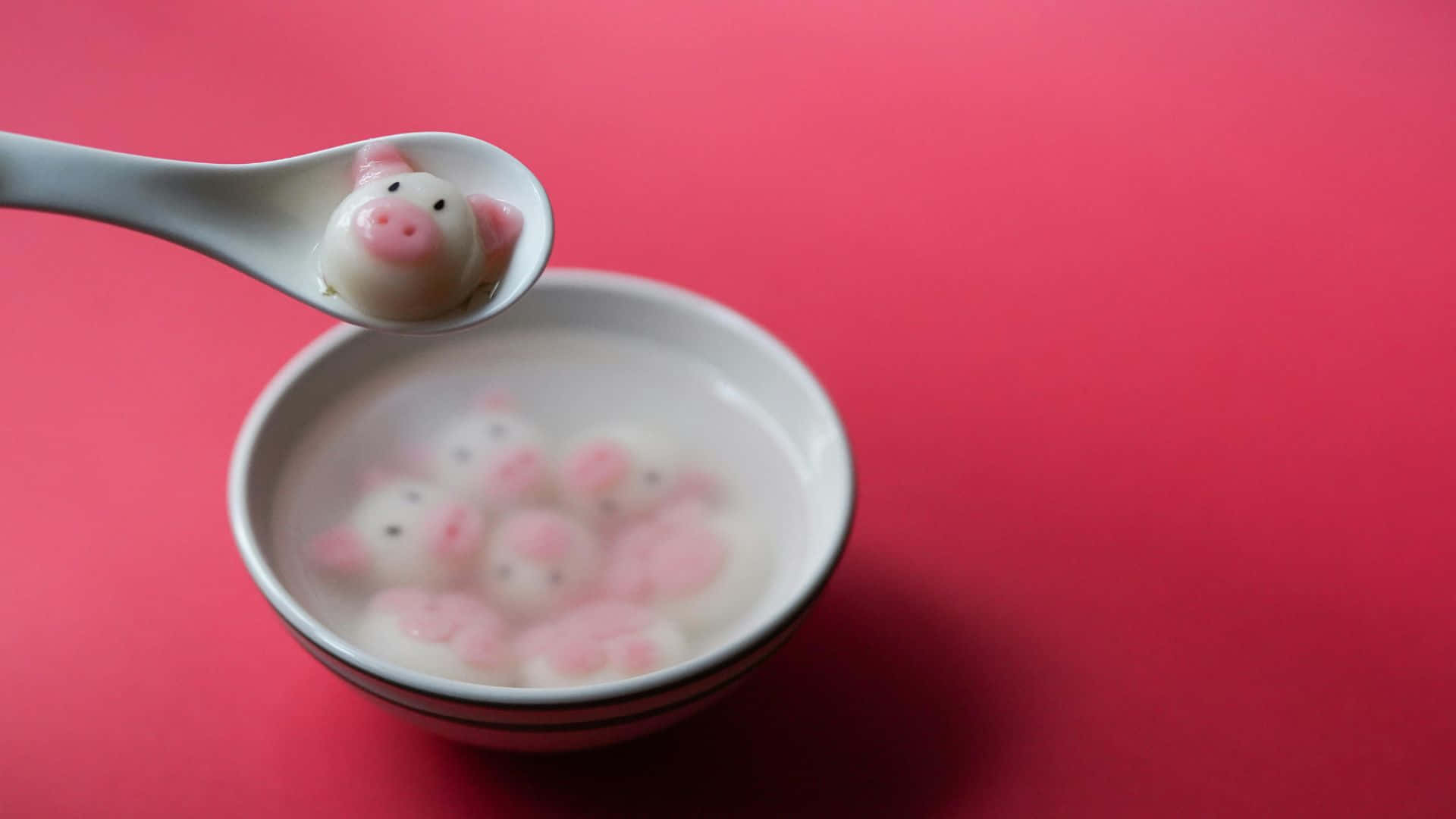  壁紙 — 一隻白色湯匙從一碗清澈的湯中舀起一個粉紅色的豬形餃子，背景是鮮豔的紅色。