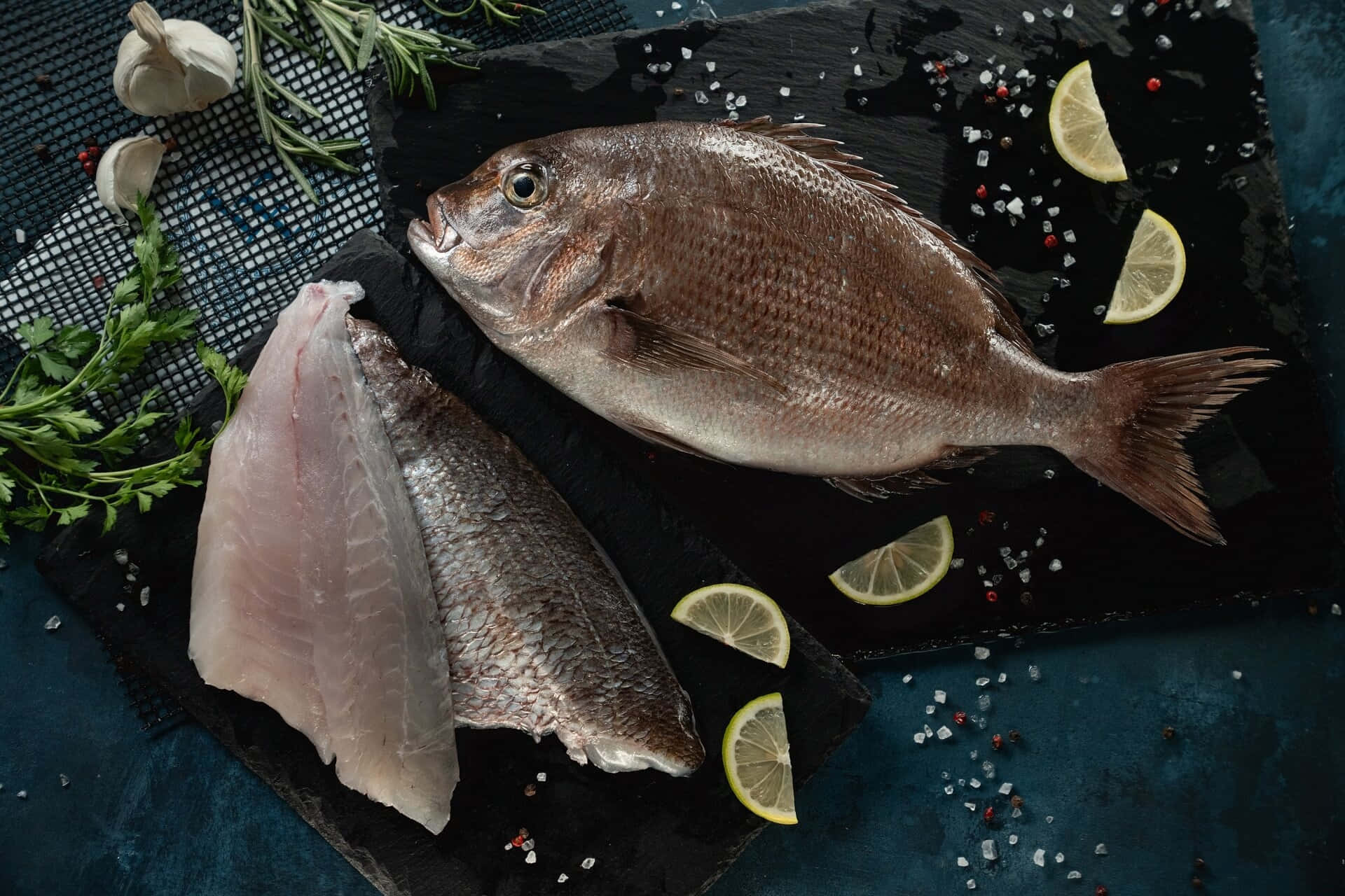 壁紙 — 一條完整的魚和魚柳放在深色石板板上，配上檸檬片、大蒜、迷迭香和鹽，營造出質樸的烹飪場景。