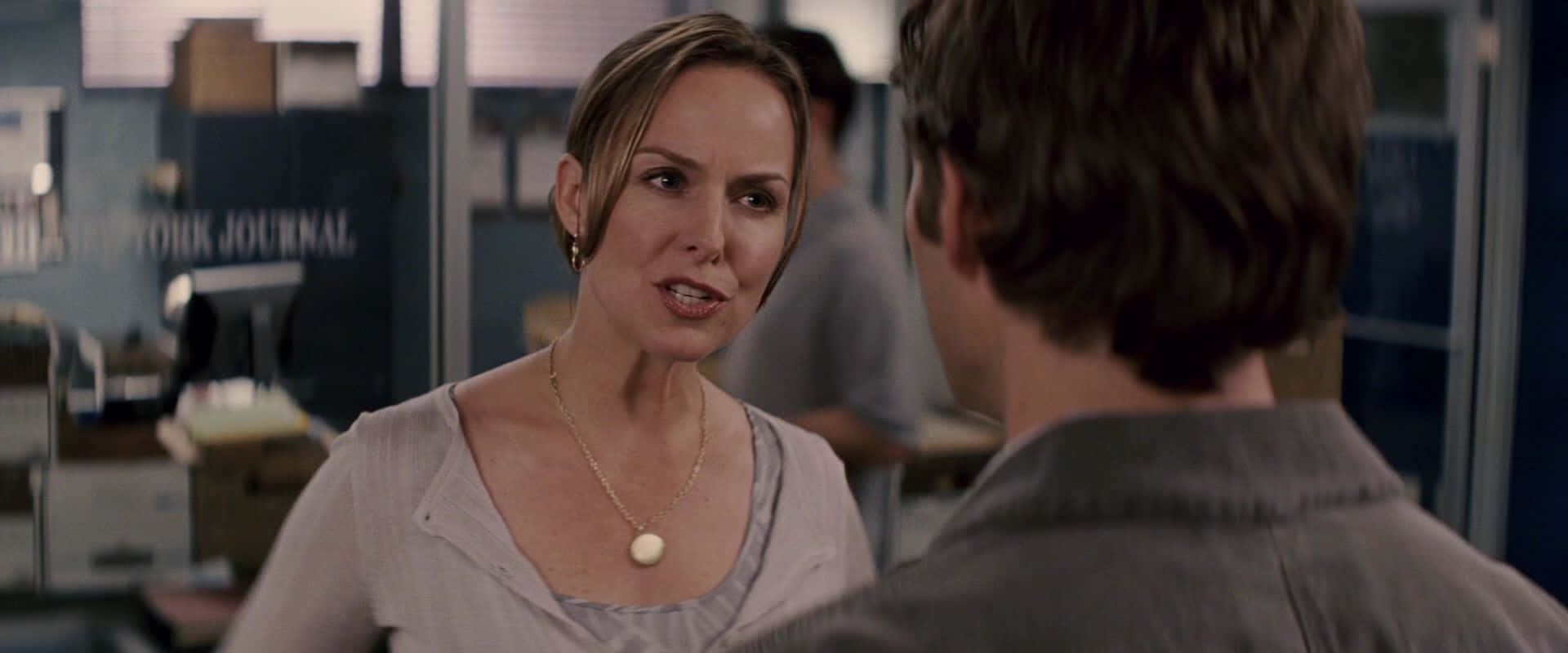 Melora Hardin 桌布 壁紙 — 一位女士在新聞編輯室裡認真地對一位穿著灰色襯衫的男士說話，背景有隔間和一個「NEW YORK JOURNAL」標誌。