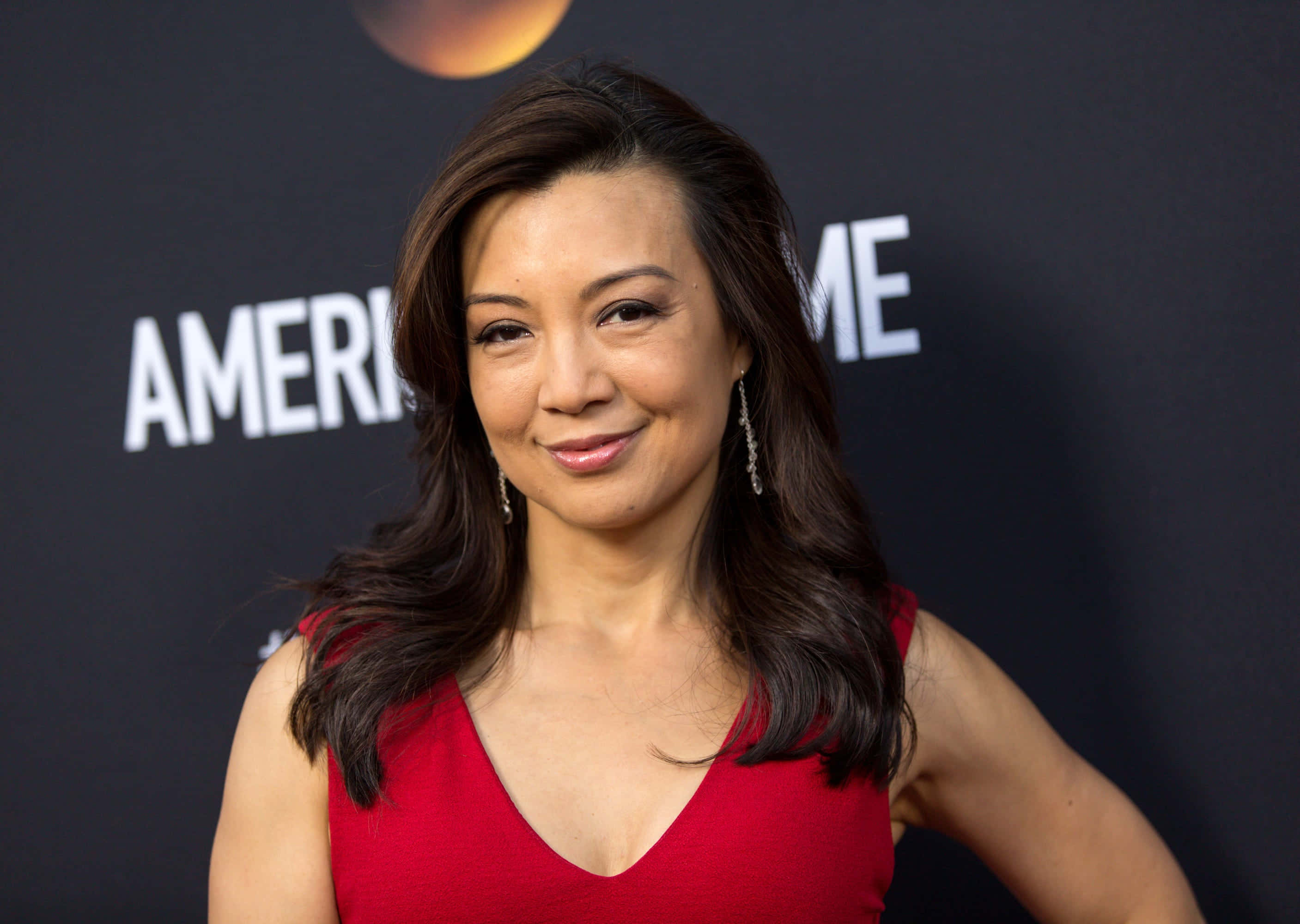 Ming Na Wen 桌布 壁紙 — 一位身穿紅色連衣裙的女性在帶有'AMERICAN'文字的深色背景前對著鏡頭微笑。