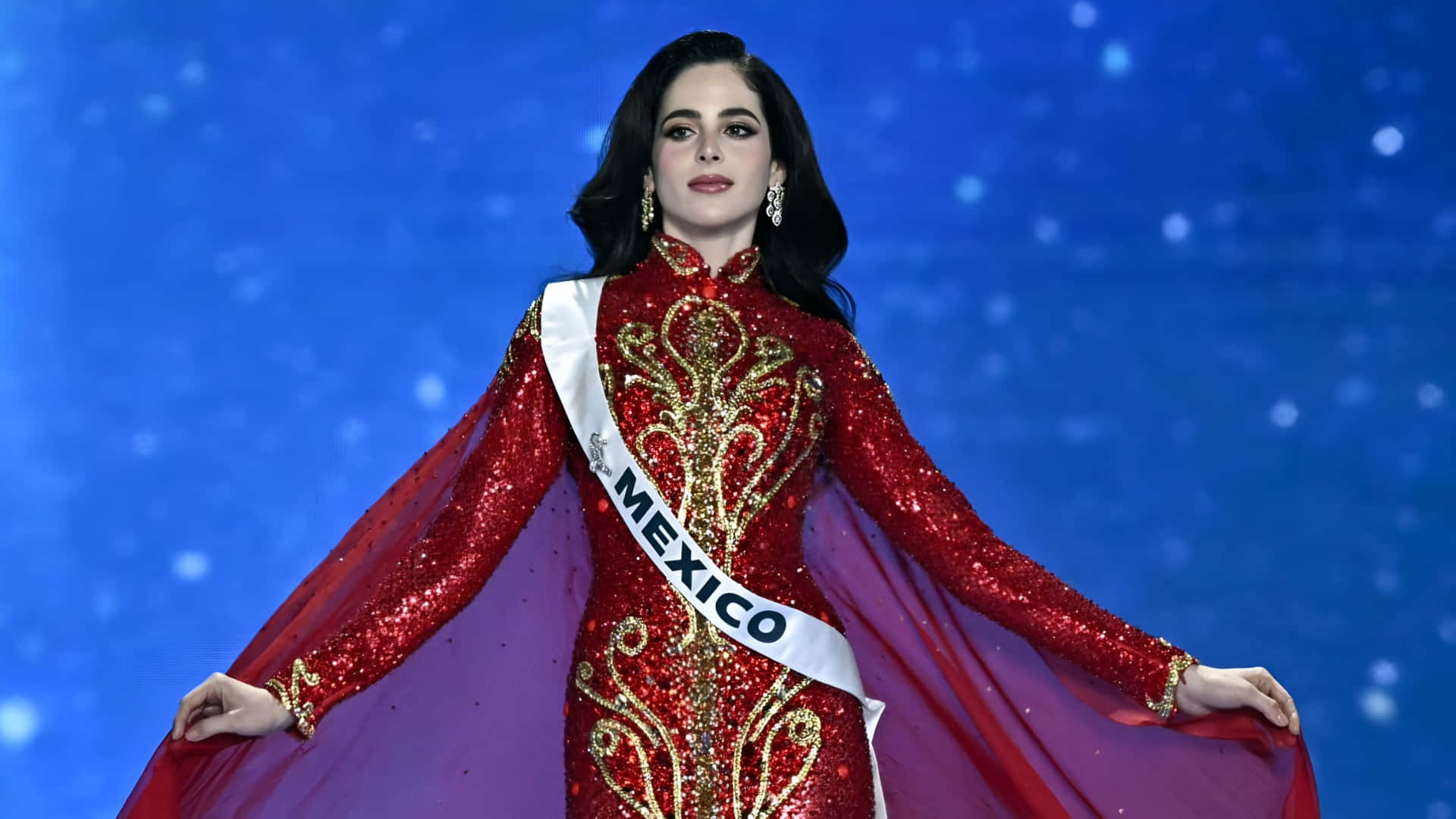  壁紙 — 一位身穿閃亮紅金色禮服、佩戴墨西哥綬帶的女性，自信地站在帶有星形燈光的藍色舞台上。