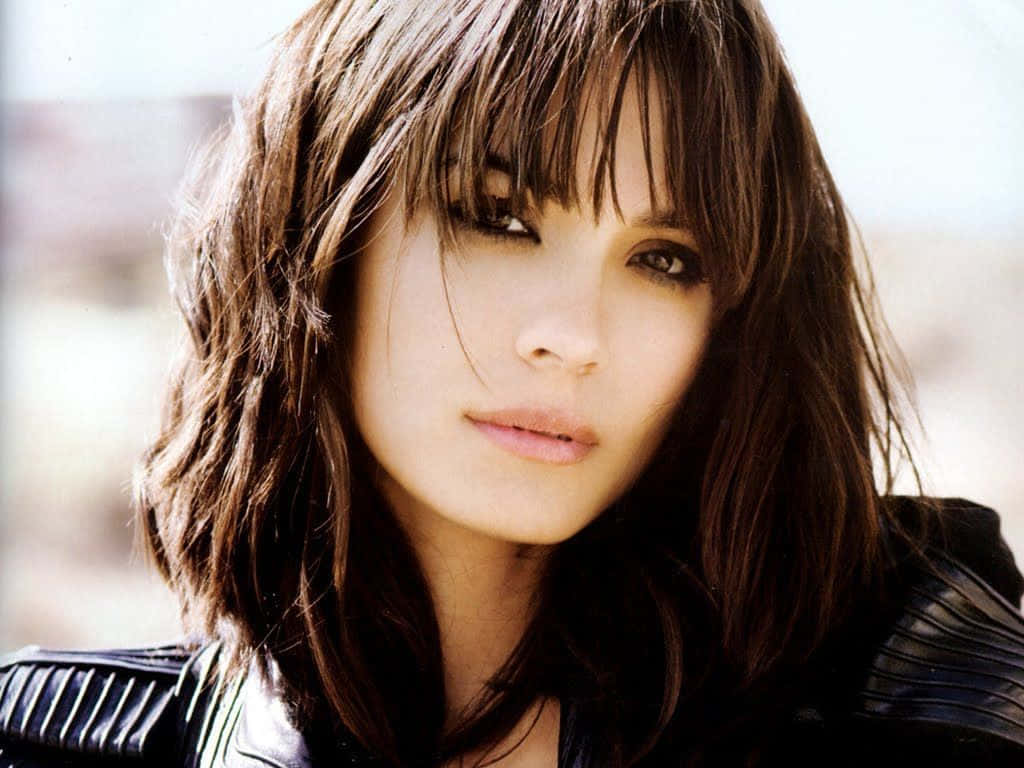 Shannyn Sossamon 桌布 壁紙 — 一位擁有深色波浪髮和瀏海的女性，在模糊的戶外背景下，穿著黑色皮夾克，溫柔地凝視著。