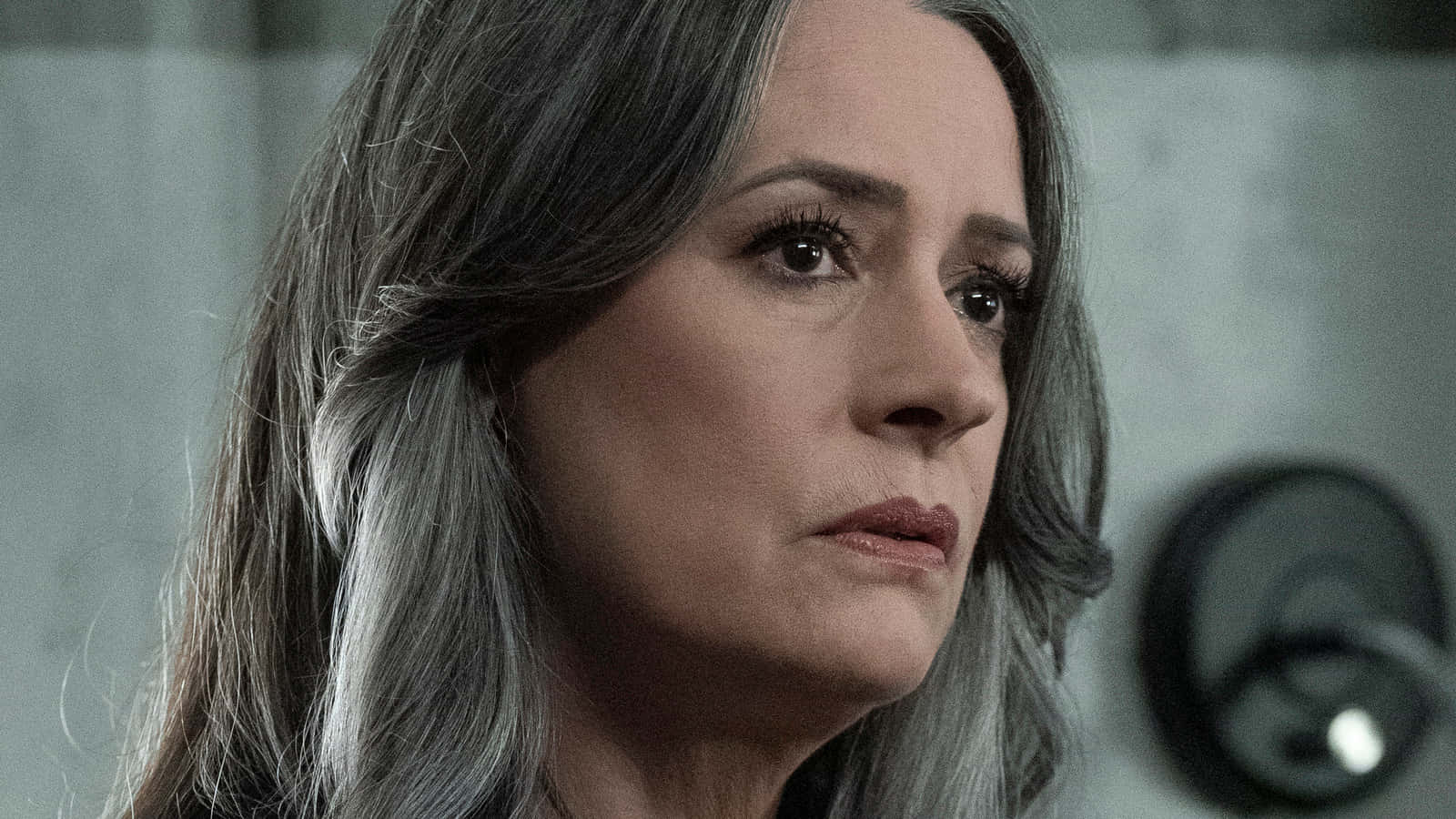 paget brewster 桌布 壁紙 — 一位灰髮女性在附近有閉路電視的昏暗混凝土房間裏若有所思地凝視着。