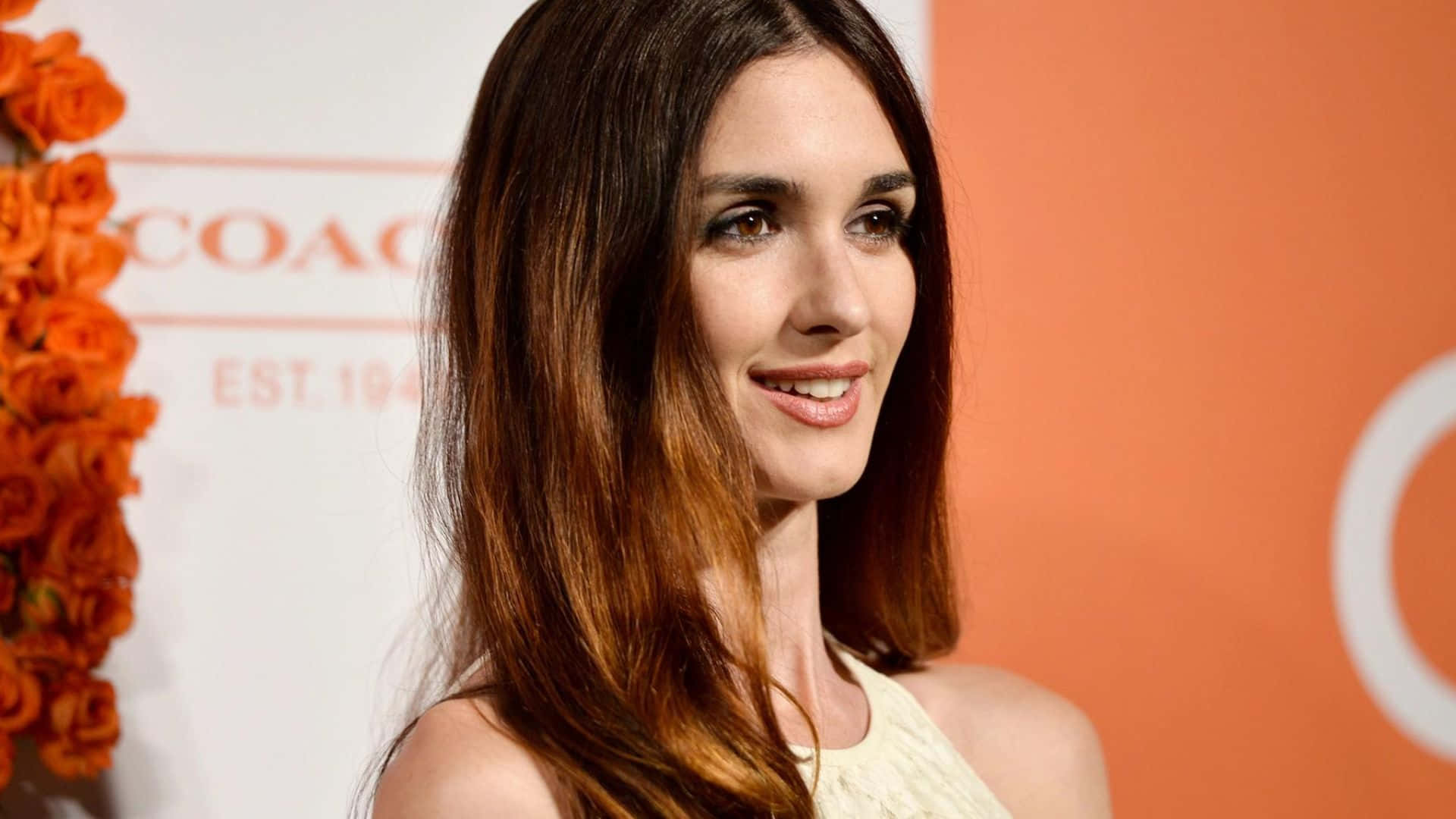 Paz Vega 桌布 壁紙 — 一位長棕色頭髮的女性在一個有橙色花卉裝飾和品牌背景的活動中溫柔微笑。
