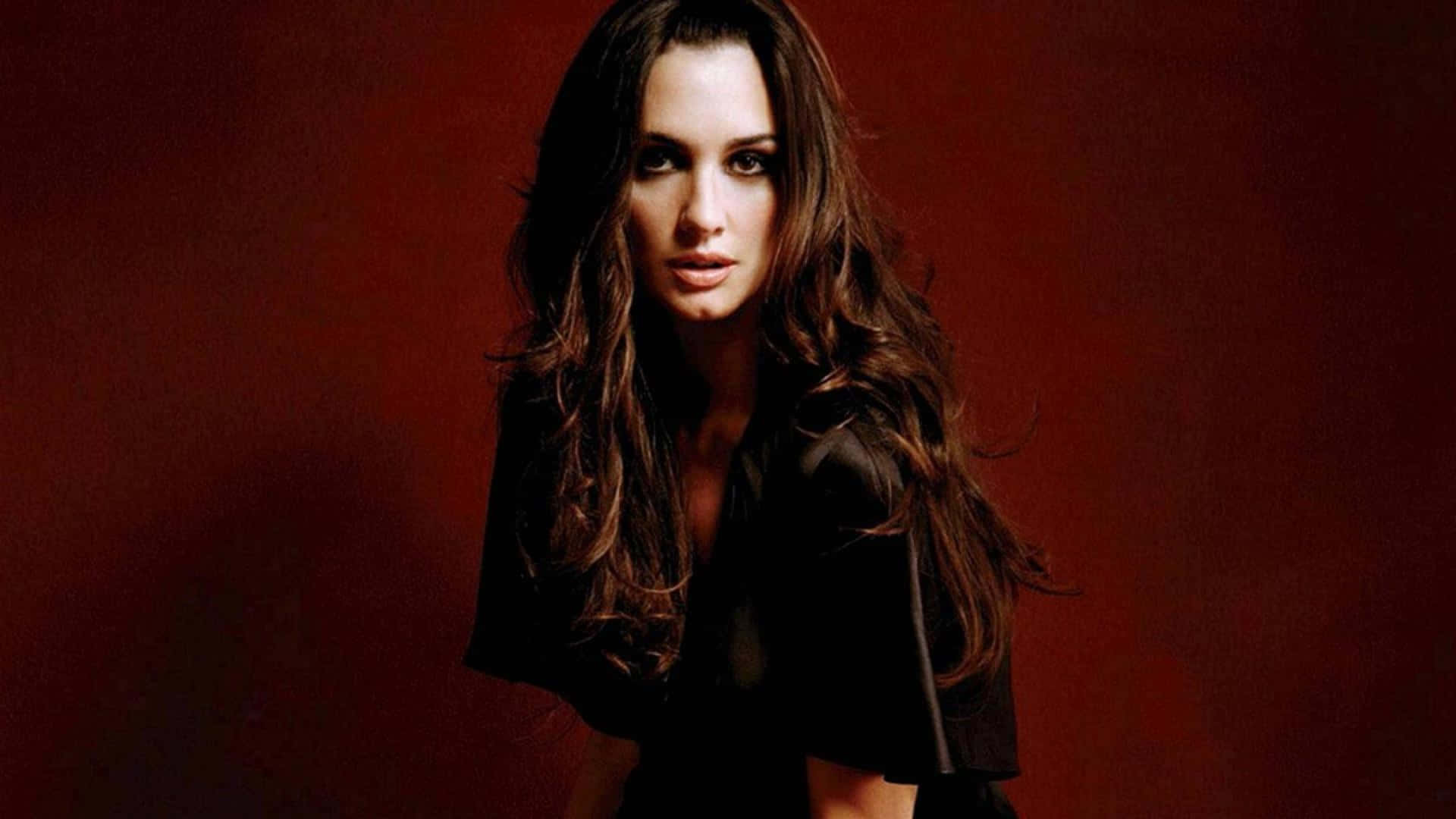Paz Vega 桌布 壁紙 — 一位長黑髮的女性，穿著黑色上衣，在深紅色工作室背景前強烈凝視。