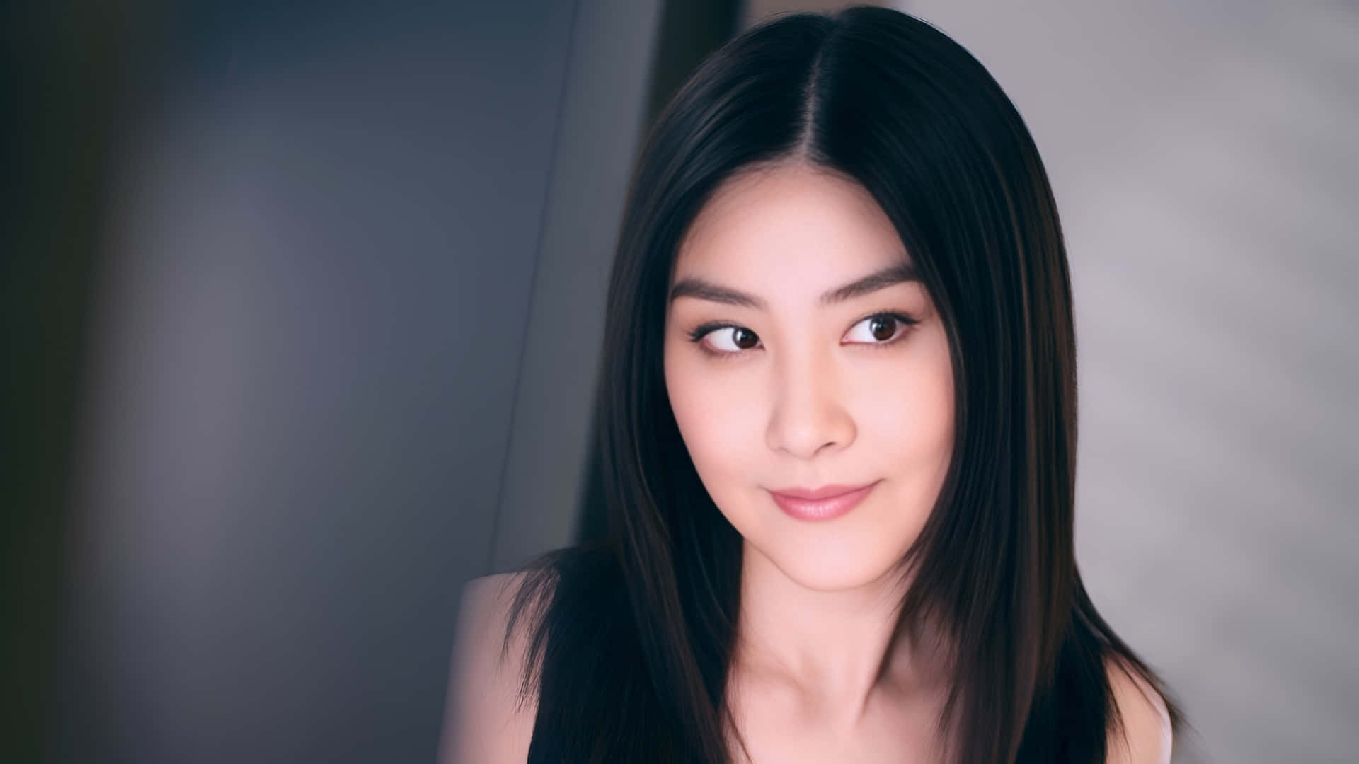  壁紙 — 一位長黑髮的女性在模糊的灰色背景前溫柔地凝視著相機。