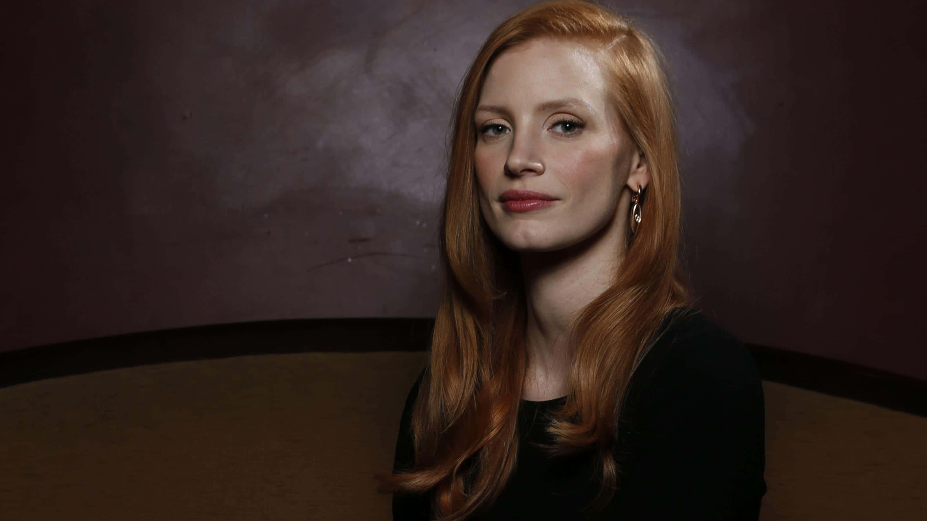 jessica chastain 桌布 壁紙 — 一位長紅髮的女性坐在昏暗的房間裡，她的臉被柔和的光線照亮，背景是深色紋理牆壁。