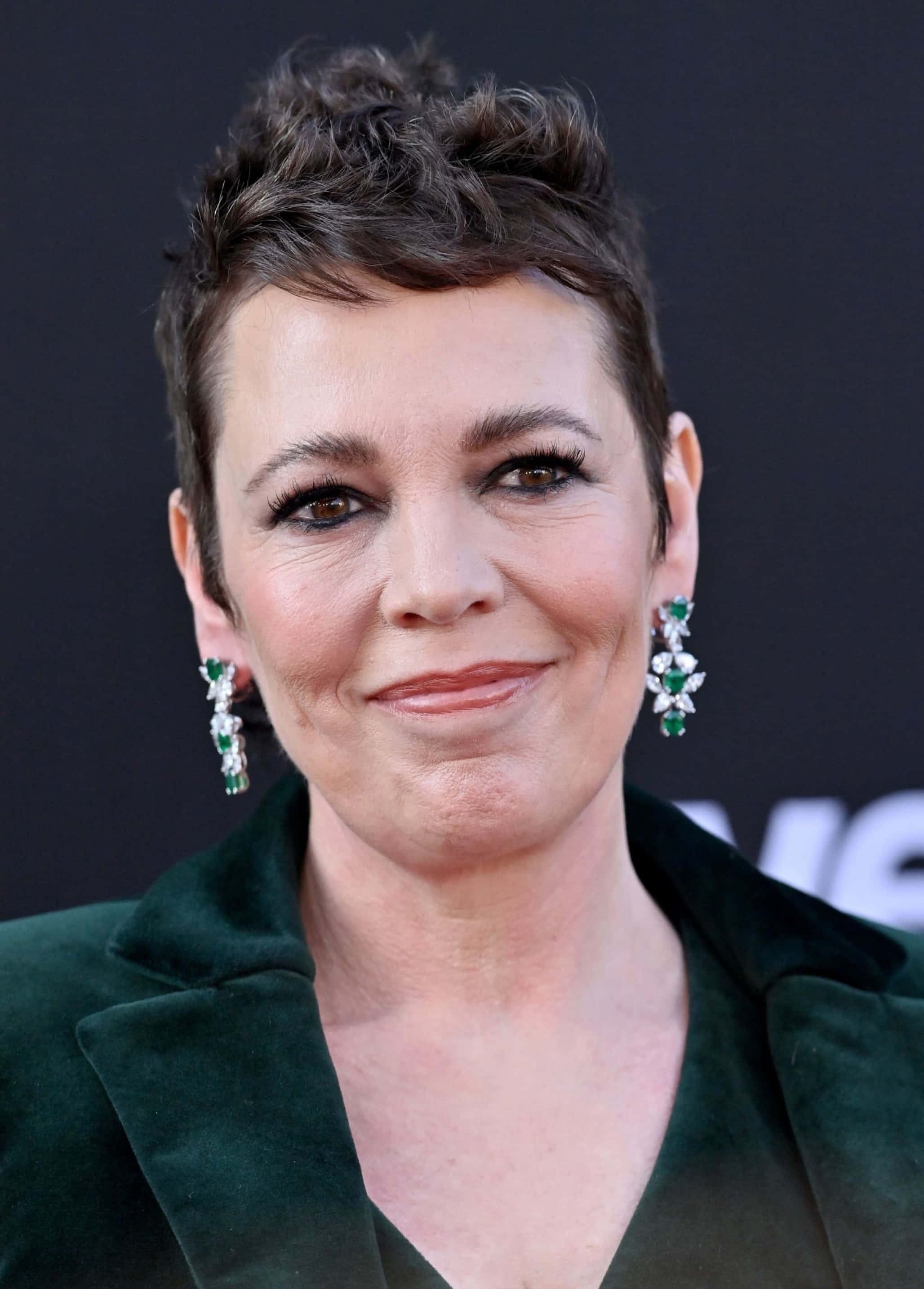 olivia colman 桌布 壁紙 — 一位短黑髮、戴綠色寶石耳環的女性，穿著深綠色天鵝絨西裝外套微笑。