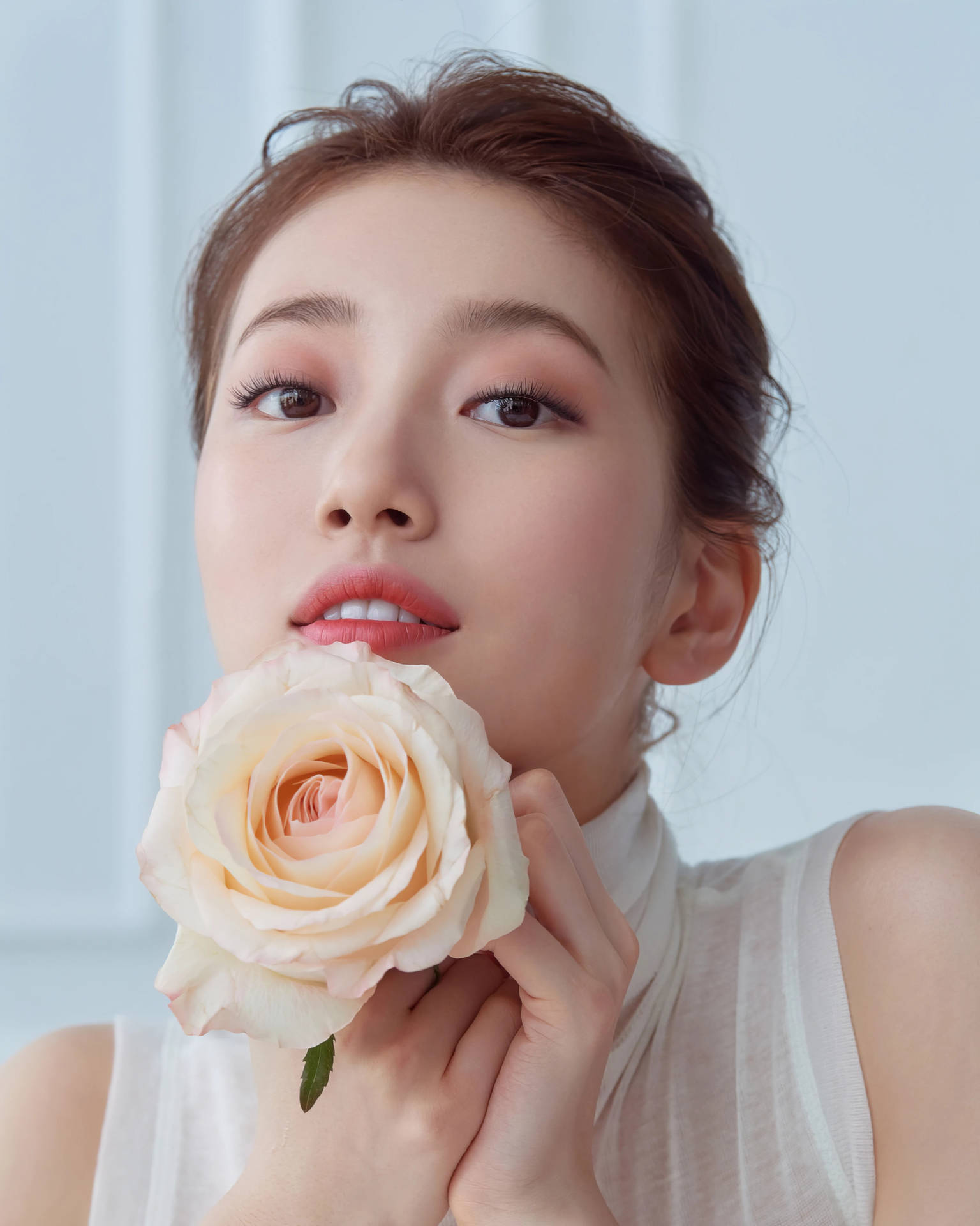 裴秀智精緻拍攝 壁紙 — 一位化著淡妝的女性，在淡藍色背景前，將一朵精緻的蜜桃玫瑰靠近嘴唇。