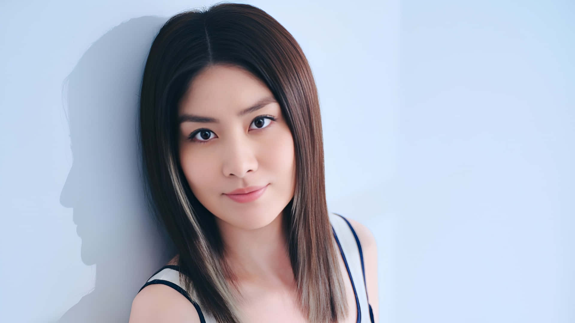  壁紙 — 一位擁有直棕色頭髮的女性靠著淺藍色牆壁，投射出柔和陰影，同時直視鏡頭。