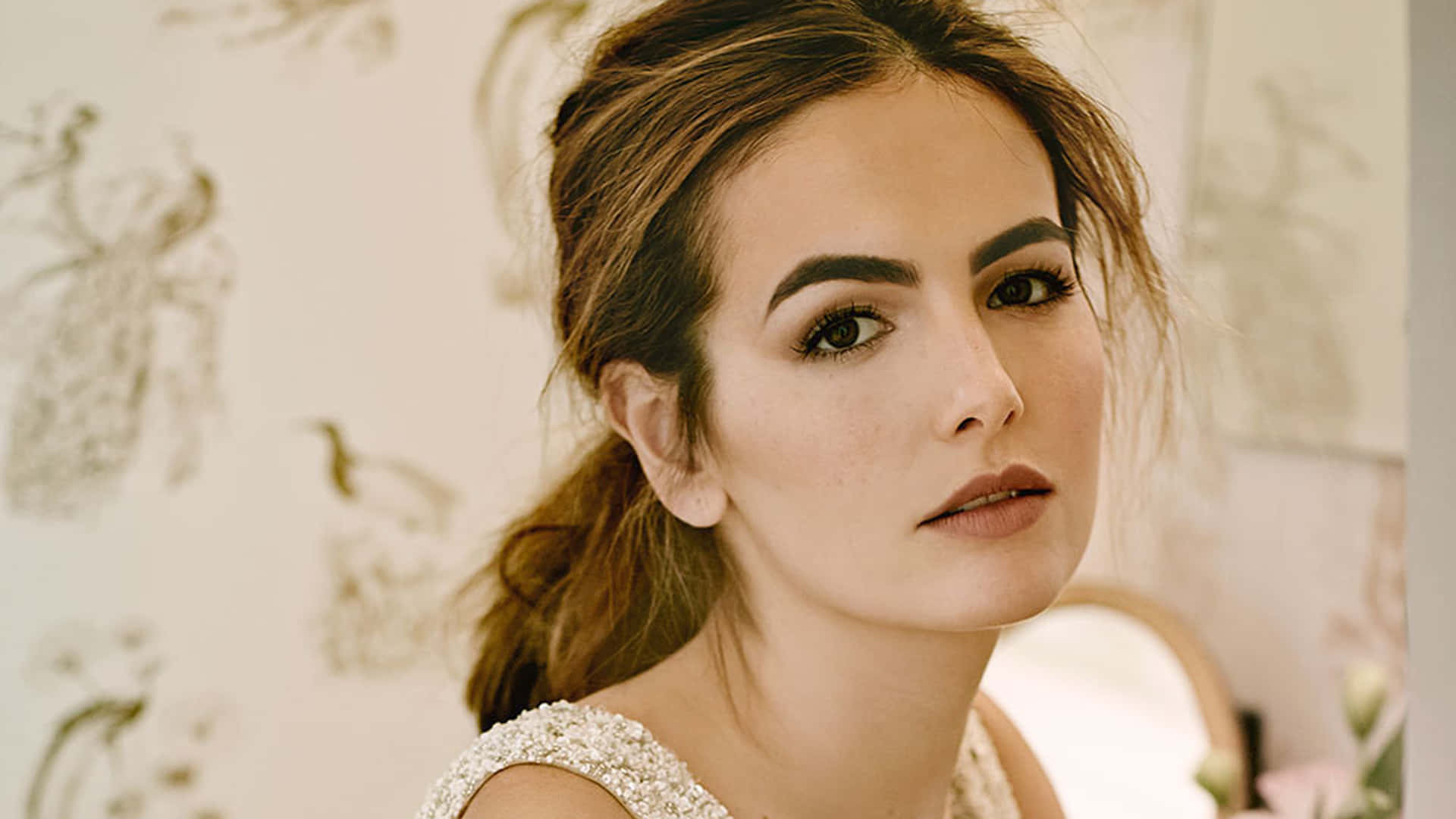 Camilla Belle 桌布 壁紙 — 一位擁有時尚髮型和優雅妝容的女性，在精緻圖案的牆紙背景下溫柔凝視。