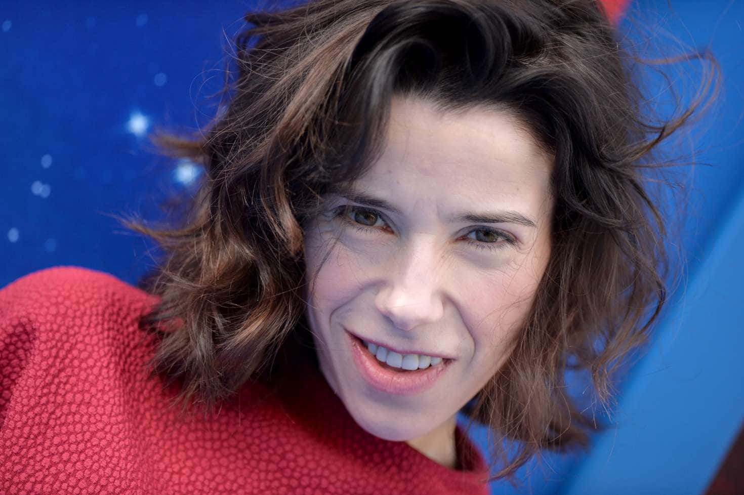 Sally Hawkins 桌布 壁紙 — 一位頭髮蓬鬆的棕髮女性，穿著質感紅色毛衣，在充滿活力的藍色星空背景下溫暖微笑。