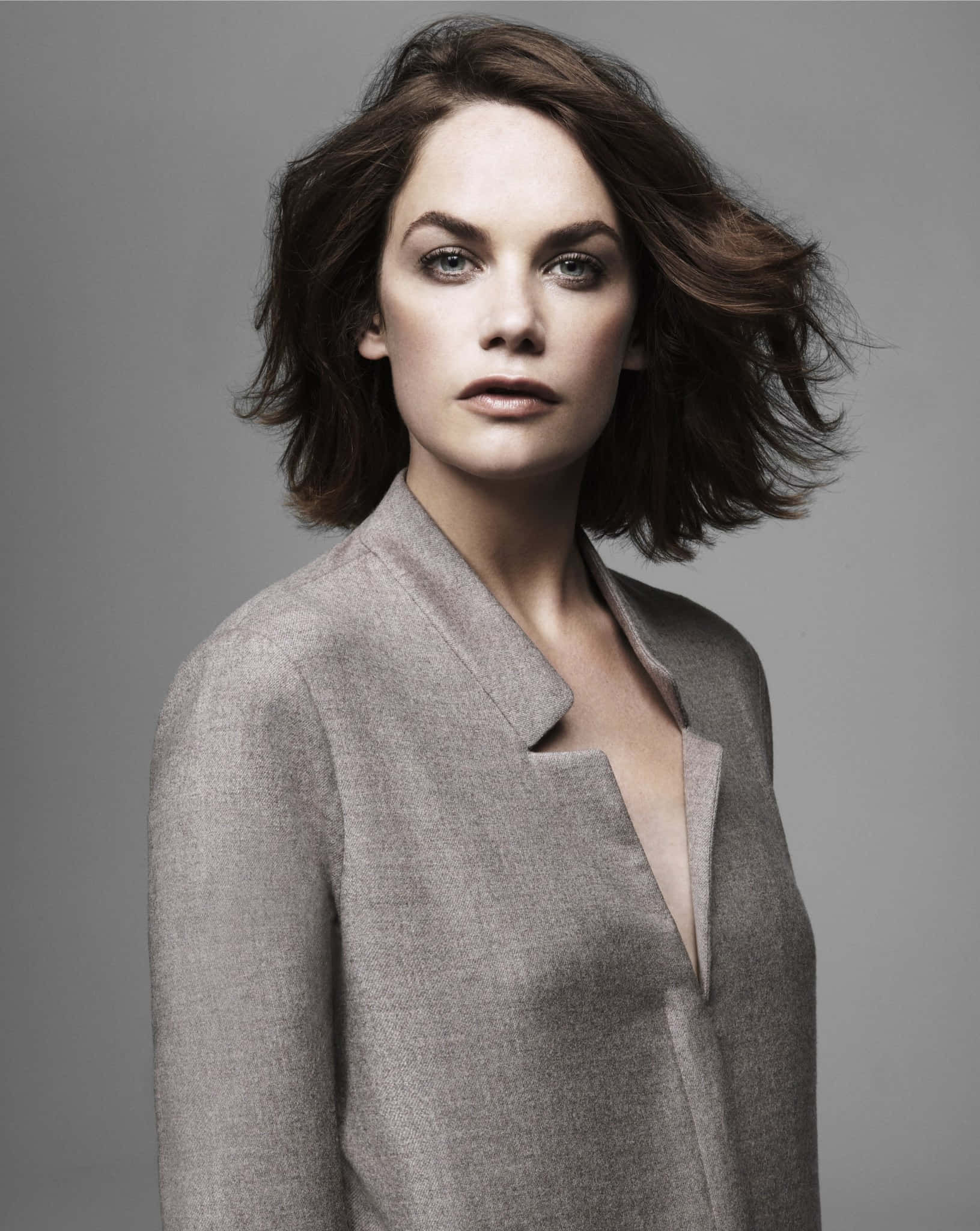 Ruth Wilson 桌布 壁紙 — 一位擁有被風吹亂的棕色頭髮和銳利眼神的女性，在純色工作室背景前穿著一件有紋理的灰色西裝外套。