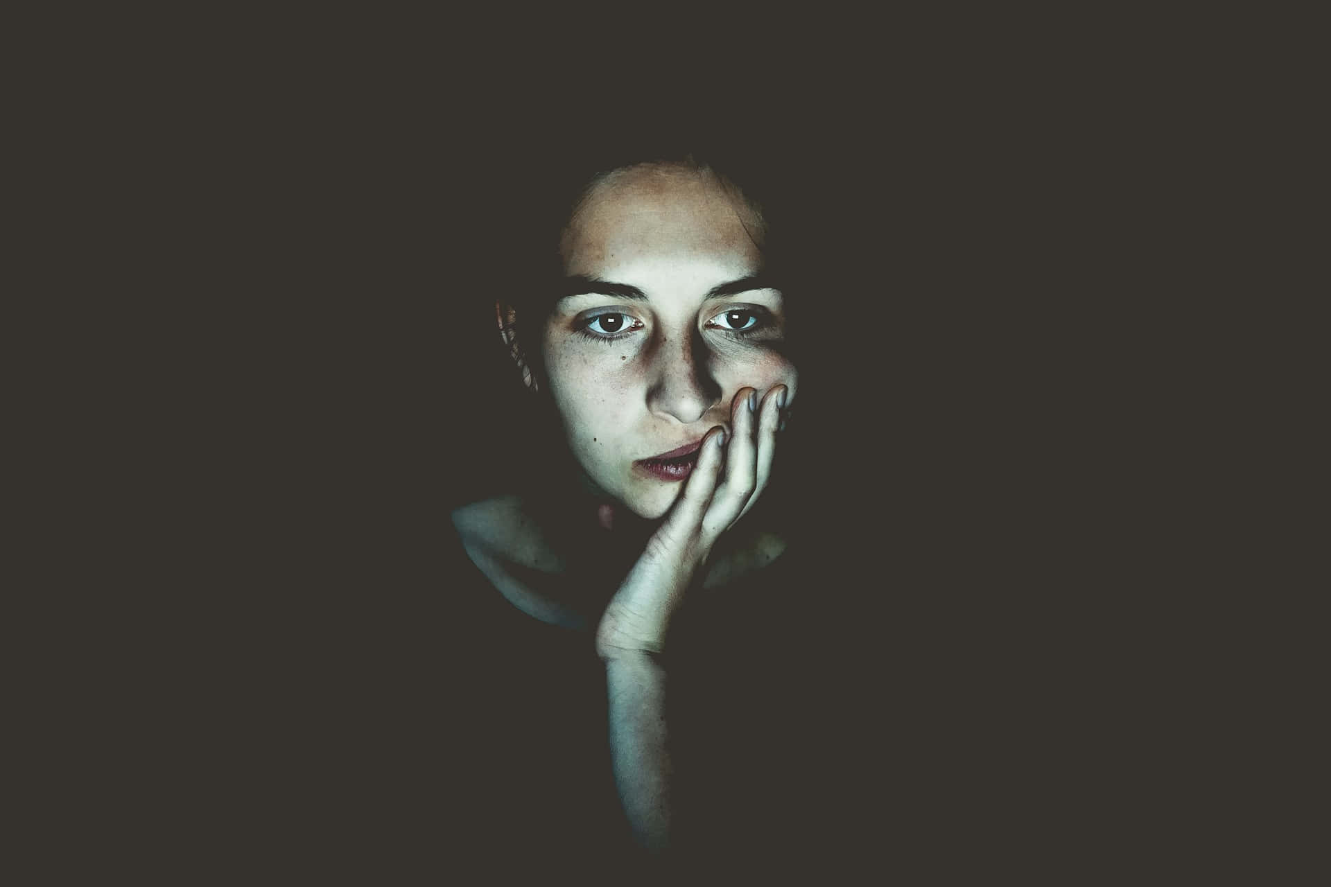  壁紙 — 一個女人的臉在黑暗中照亮，手放在嘴邊，眼睛直視，表情憂鬱。