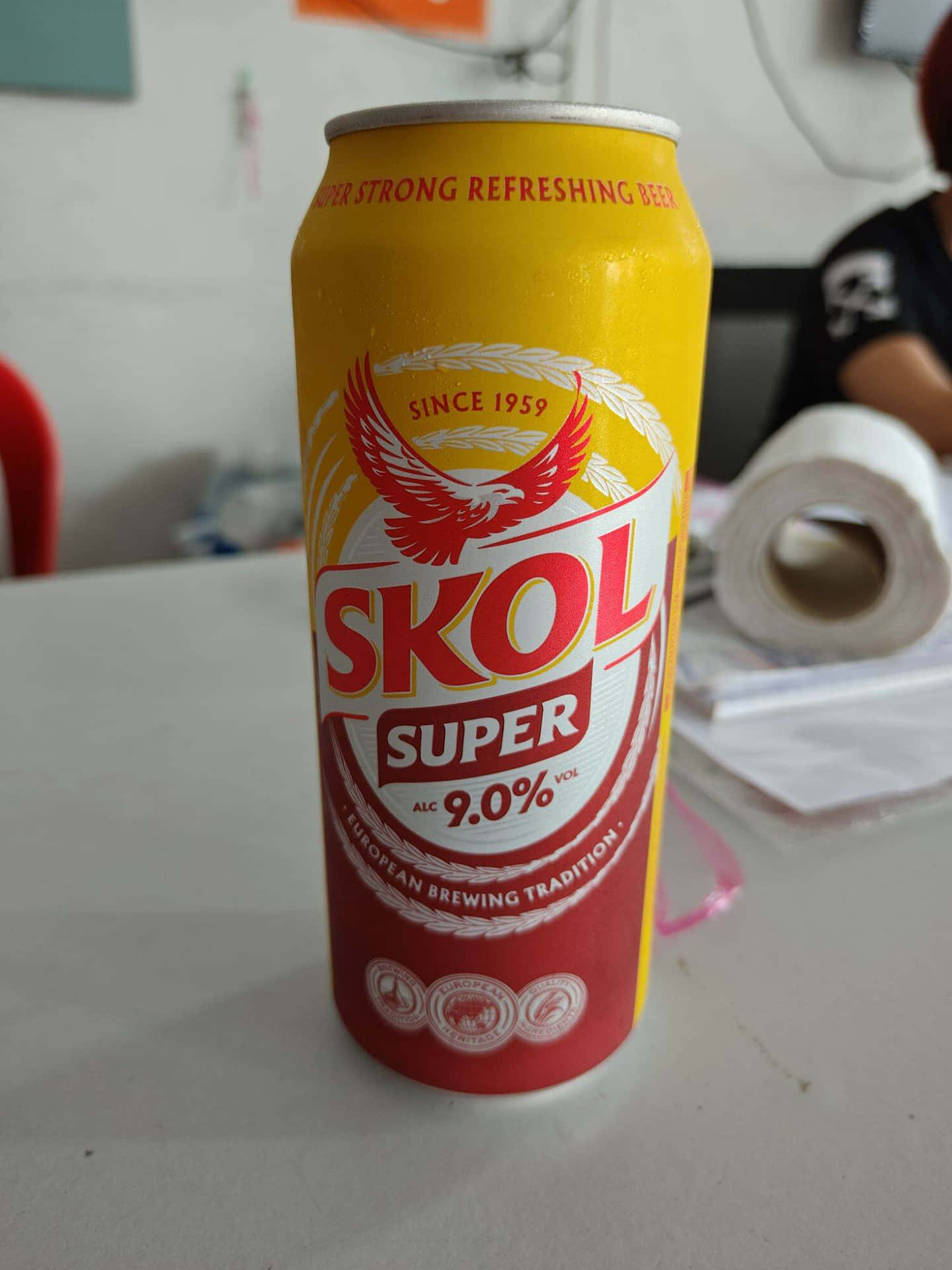  壁紙 — 一個黃色和紅色的Skol Super啤酒罐，酒精含量9.0%，放在白色桌子上，背景模糊。